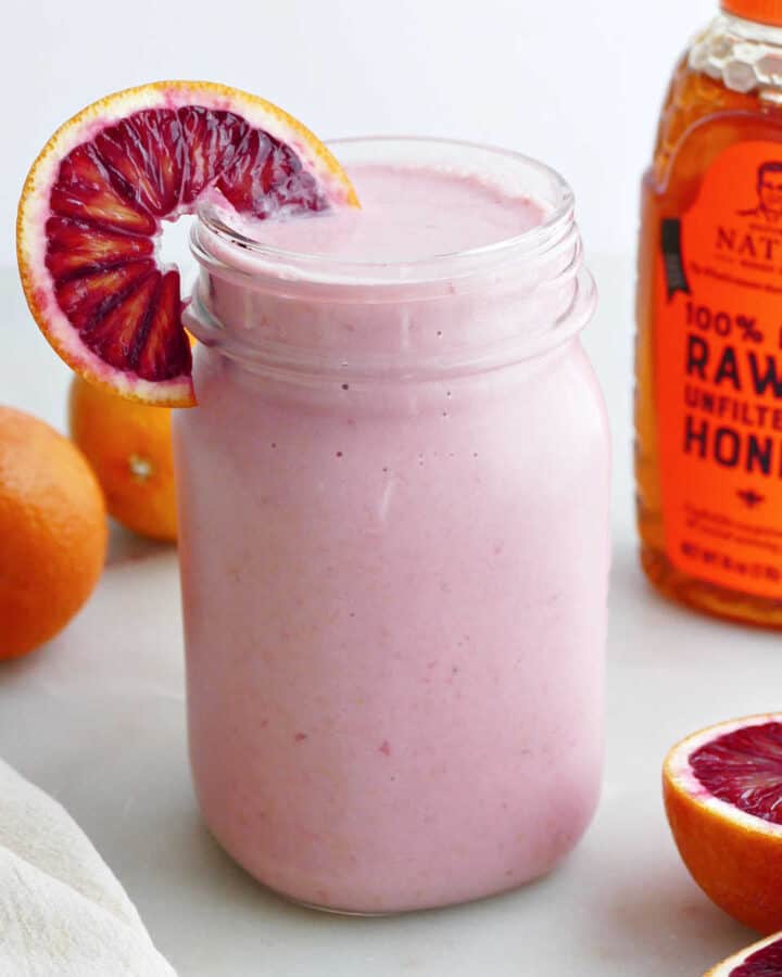 blood orange smoothie in a glass with an orange wedge in front of honey bottle