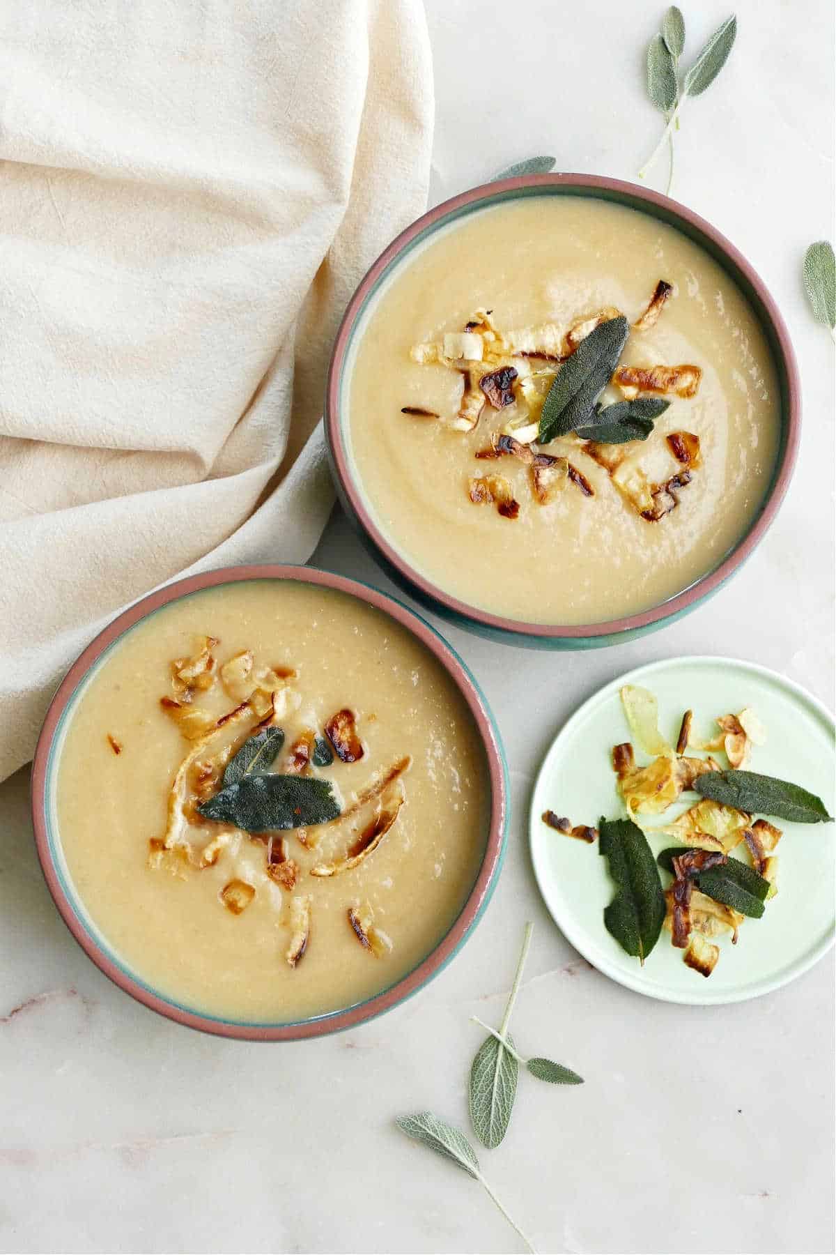 two bowls of celeriac and parsnip soup topped with fried sage on a counter