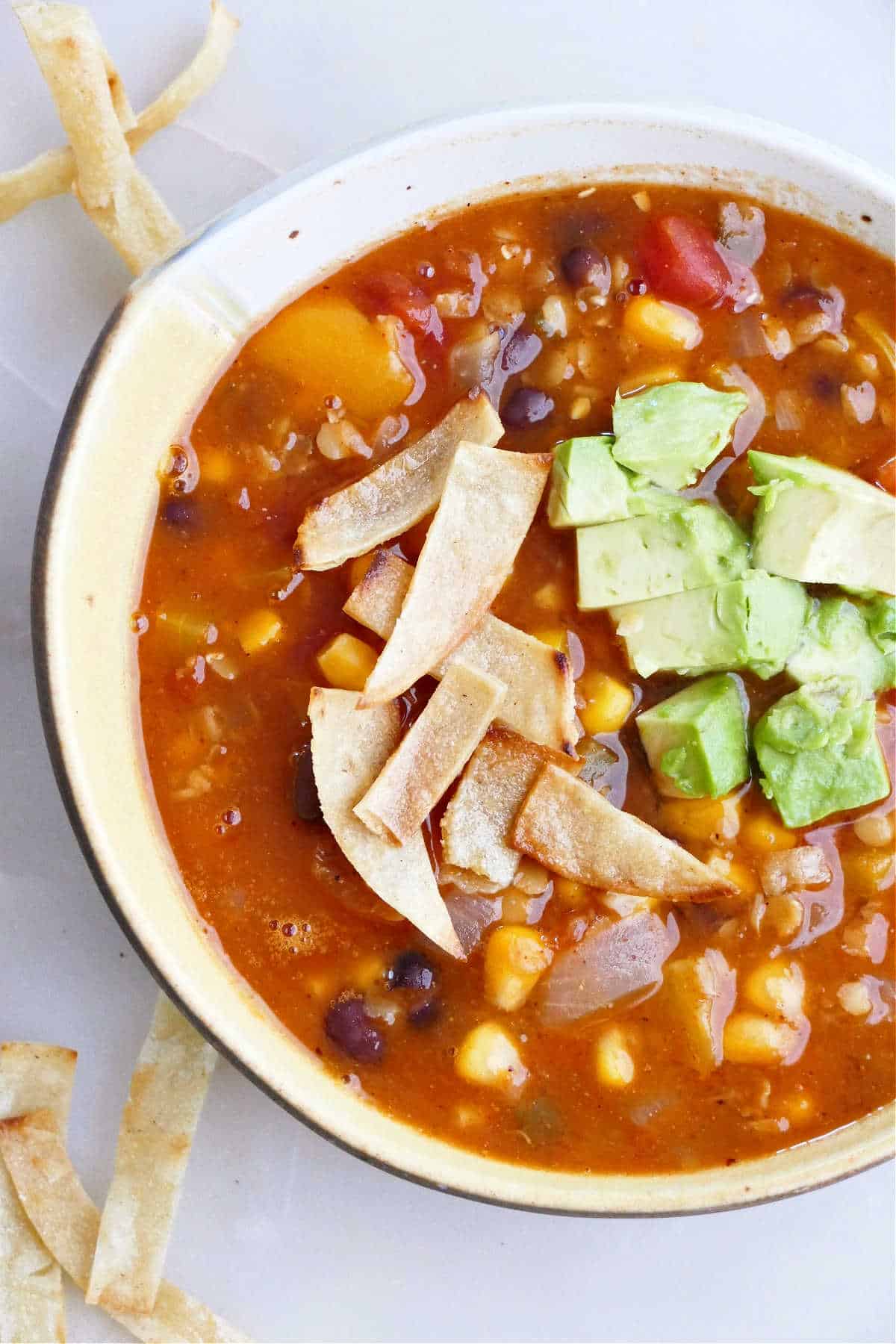 bowl of red lentil tortilla soup topped with crispy tortilla strips and avocados on a counter