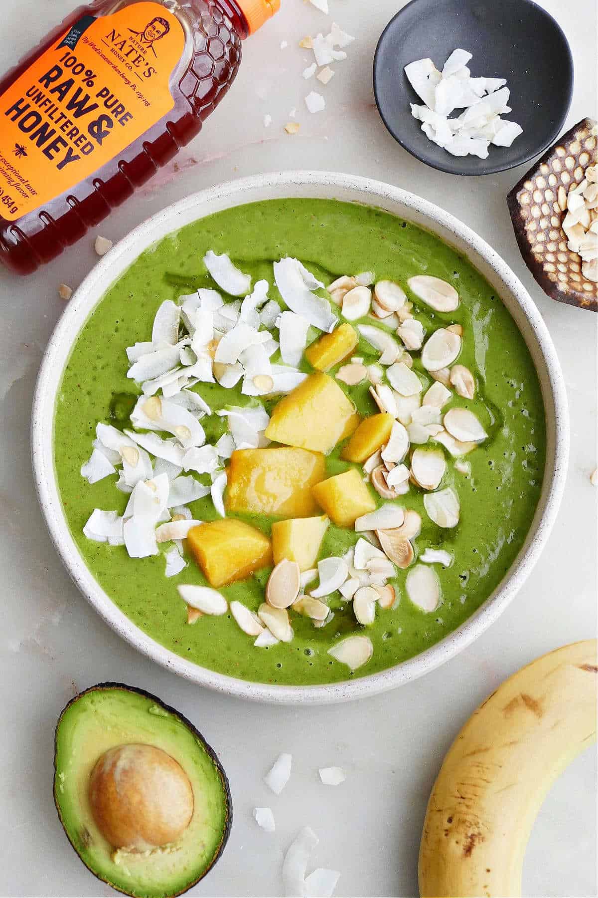 mango kale smoothie bowl topped with coconut, mango, and almonds surrounded by ingredients