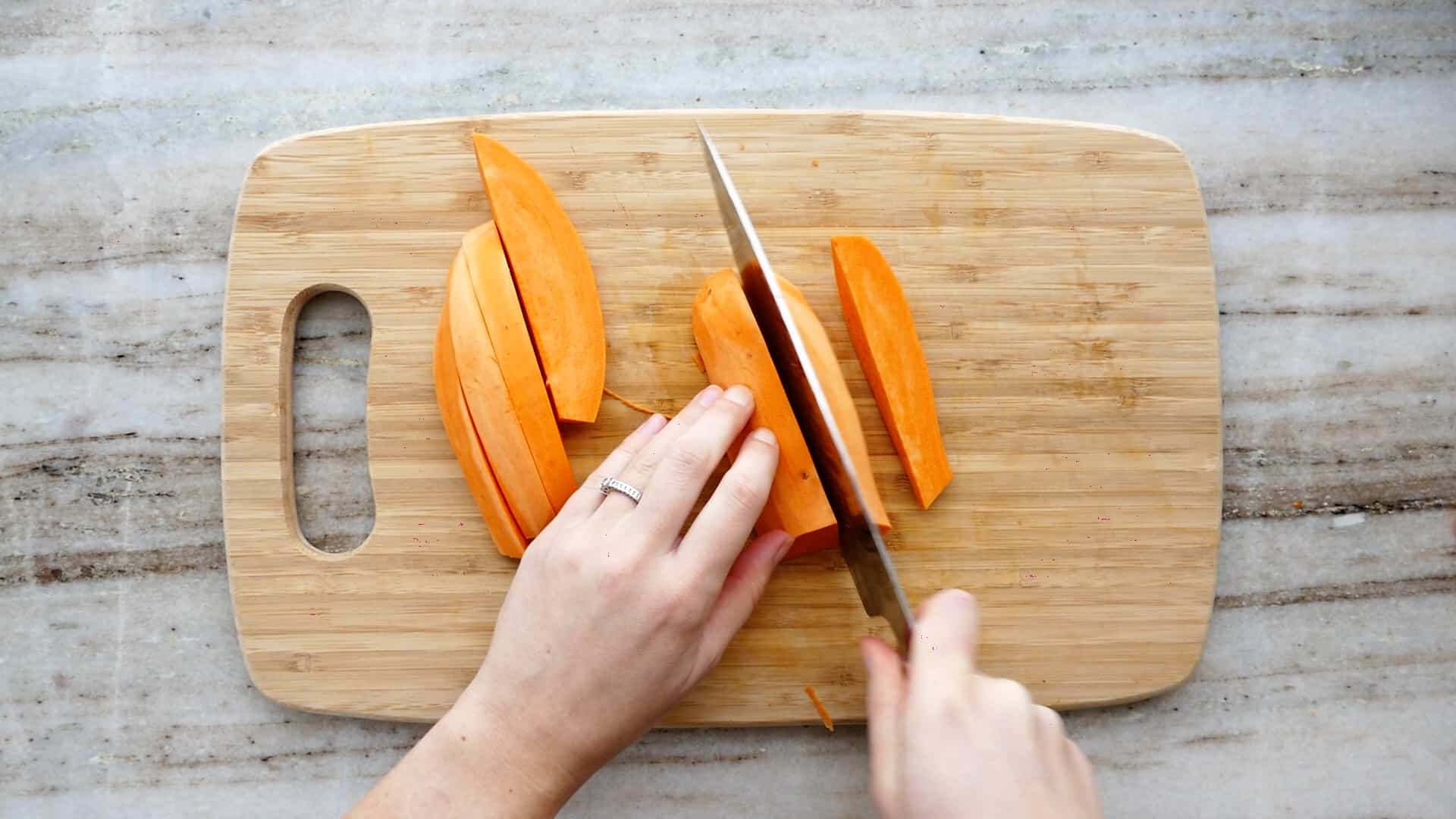 woman cutting sweet potatoes into wedges with a knife and bamboo cutting board