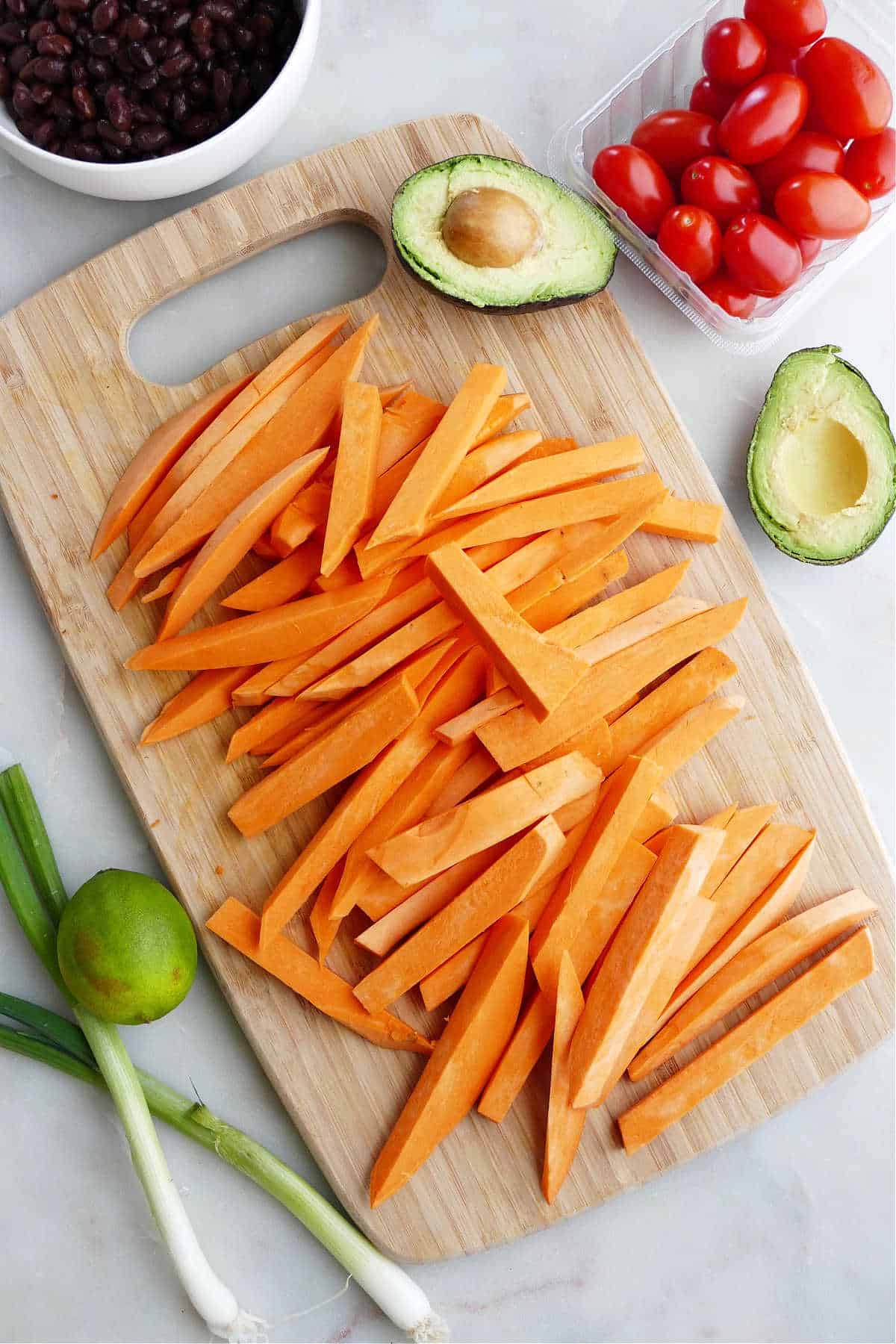 sweet potato sliced into fries on a cutting board next to avocado, tomatoes, lime, and onions