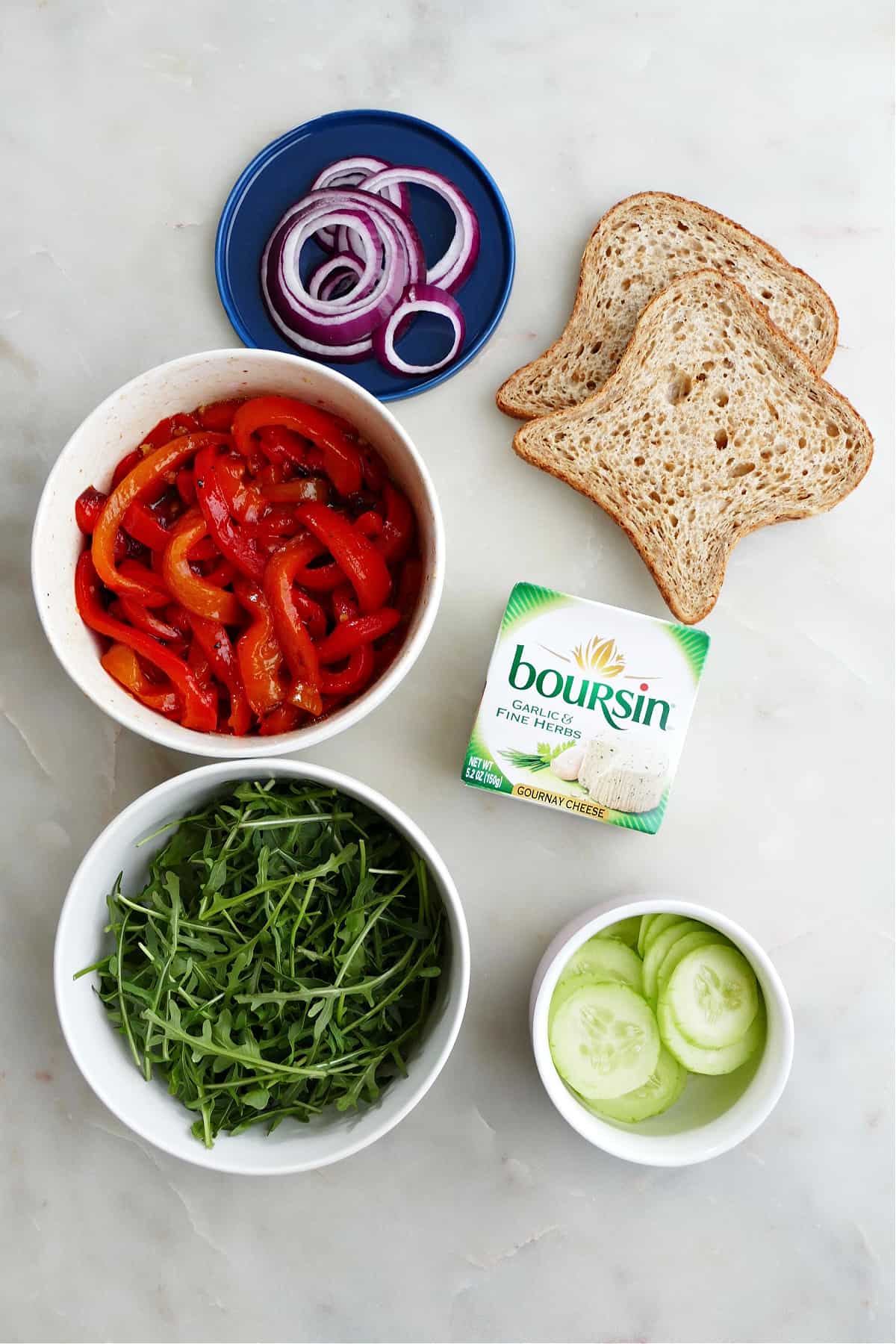 red onion, red peppers, arugula, cucumber, Boursin cheese, and sliced bread on a counter
