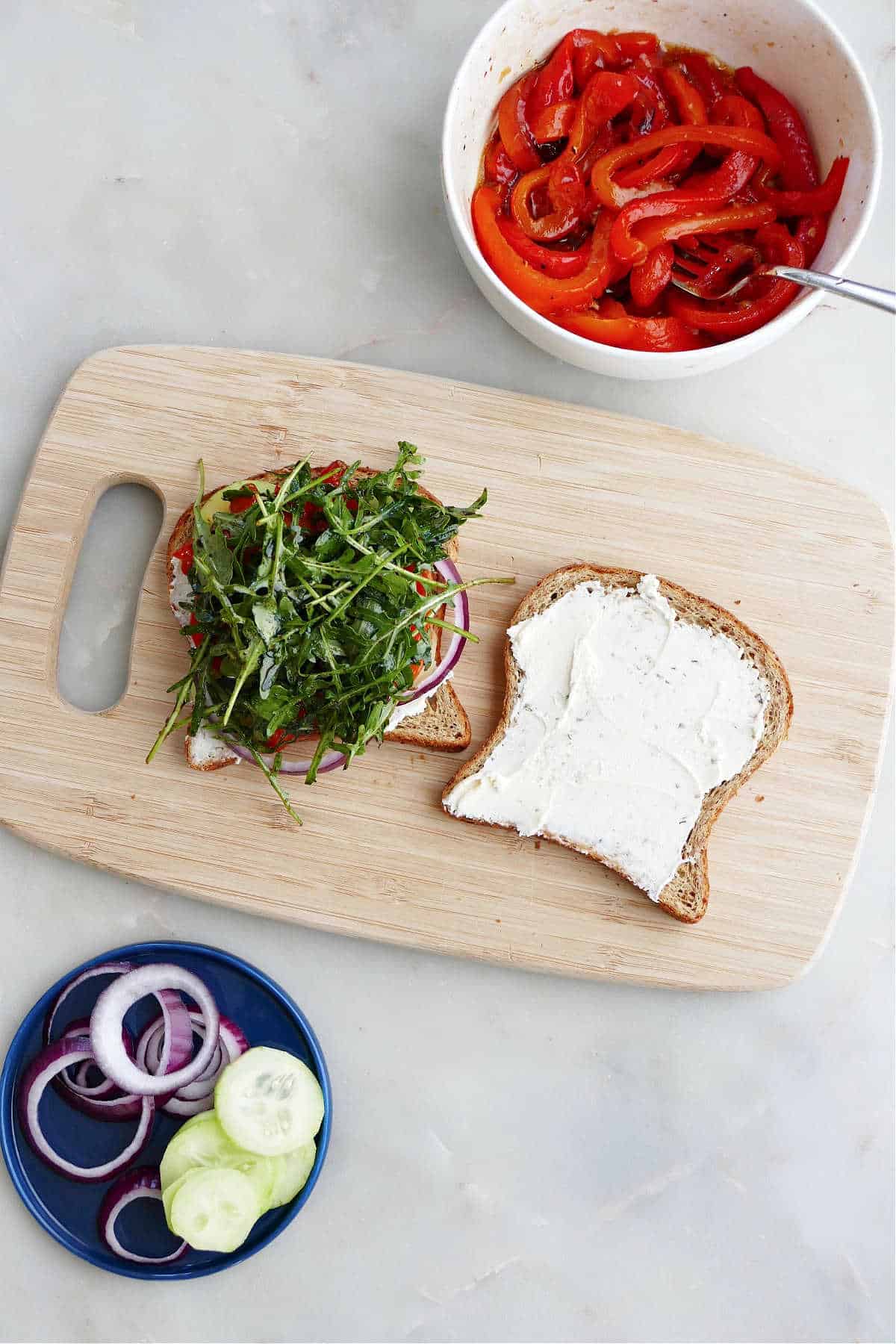 a vegetable cheese sandwich being prepared on a cutting board next to bowls of toppings