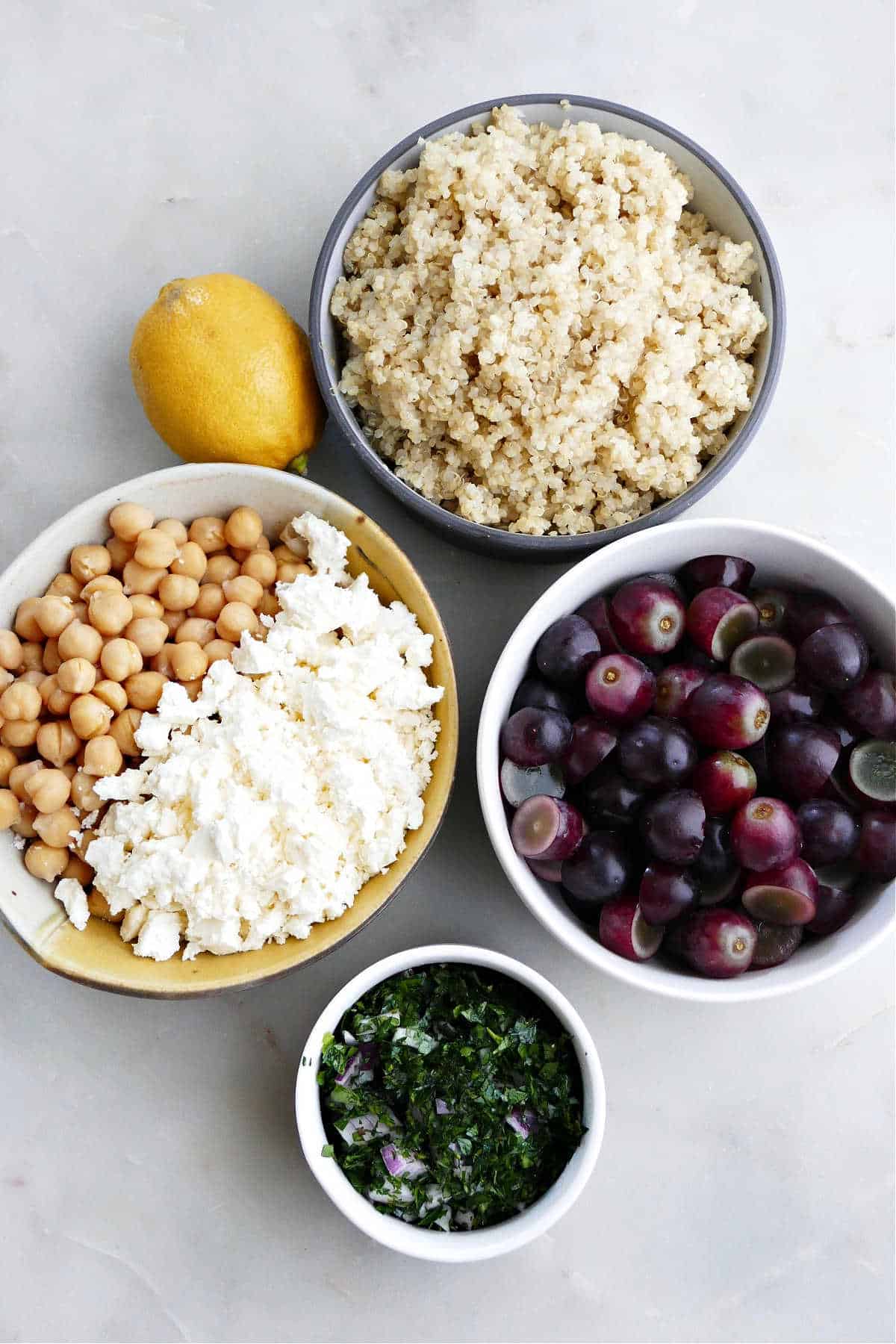 quinoa, grapes, chopped herbs, feta cheese, chickpeas, and a lemon on a counter