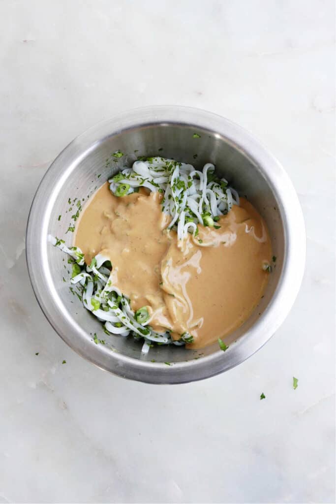 rice noodles, scallions, herbs, and a creamy peanut sauce in a mixing bowl on a counter