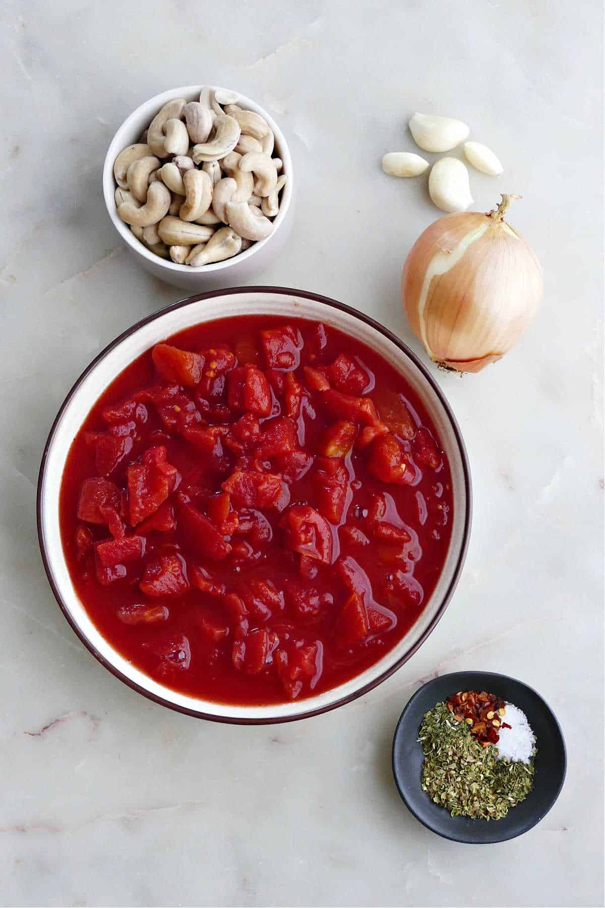 canned tomatoes, cashews, garlic cloves, onion, and seasonings spread out on a counter