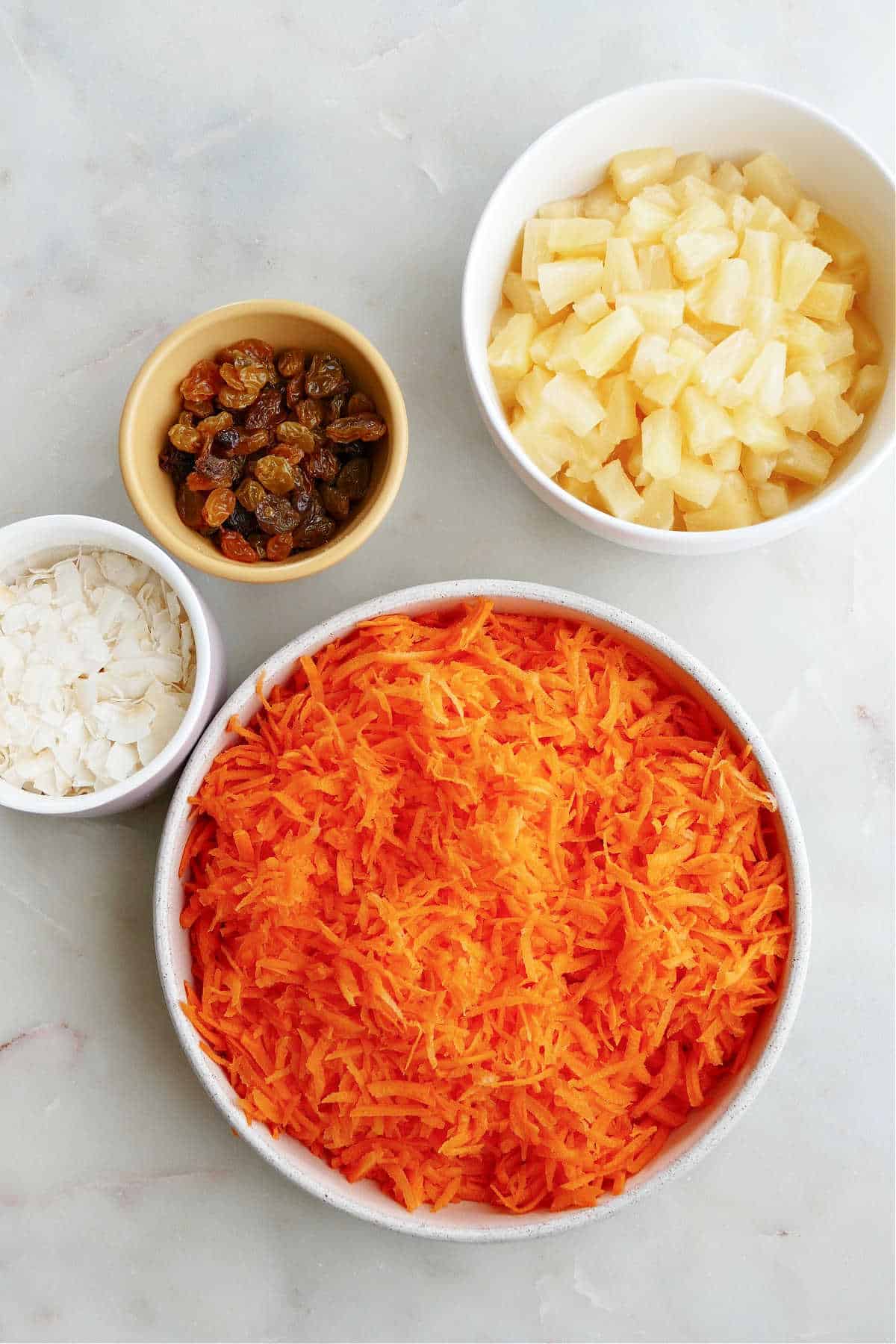 bowls of grated carrots, canned pineapple, coconut flakes, and golden raisins on a counter