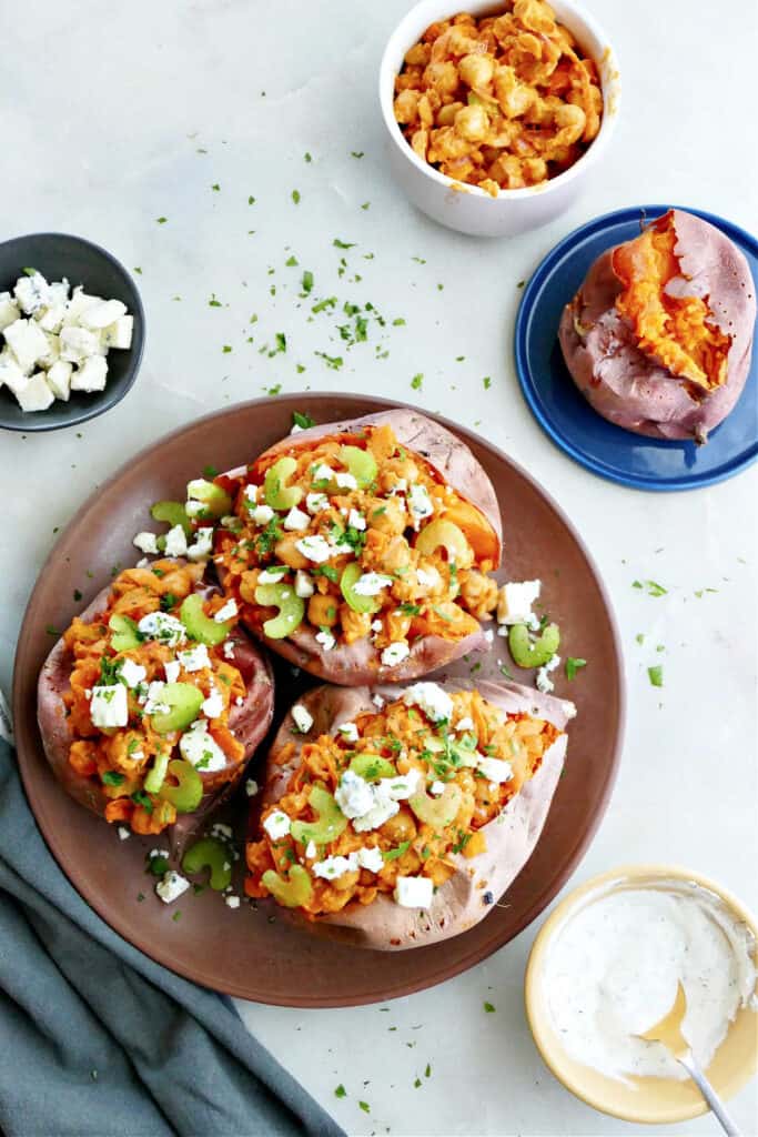 three buffalo chickpea stuffed sweet potatoes on a serving plate next to plates with toppings