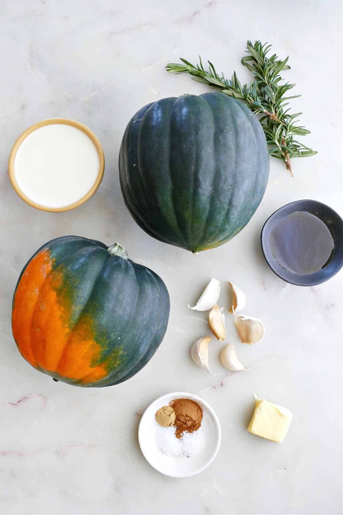 two acorn squash, rosemary sprigs, cream, garlic cloves, maple syrup, spices, and butter on a counter