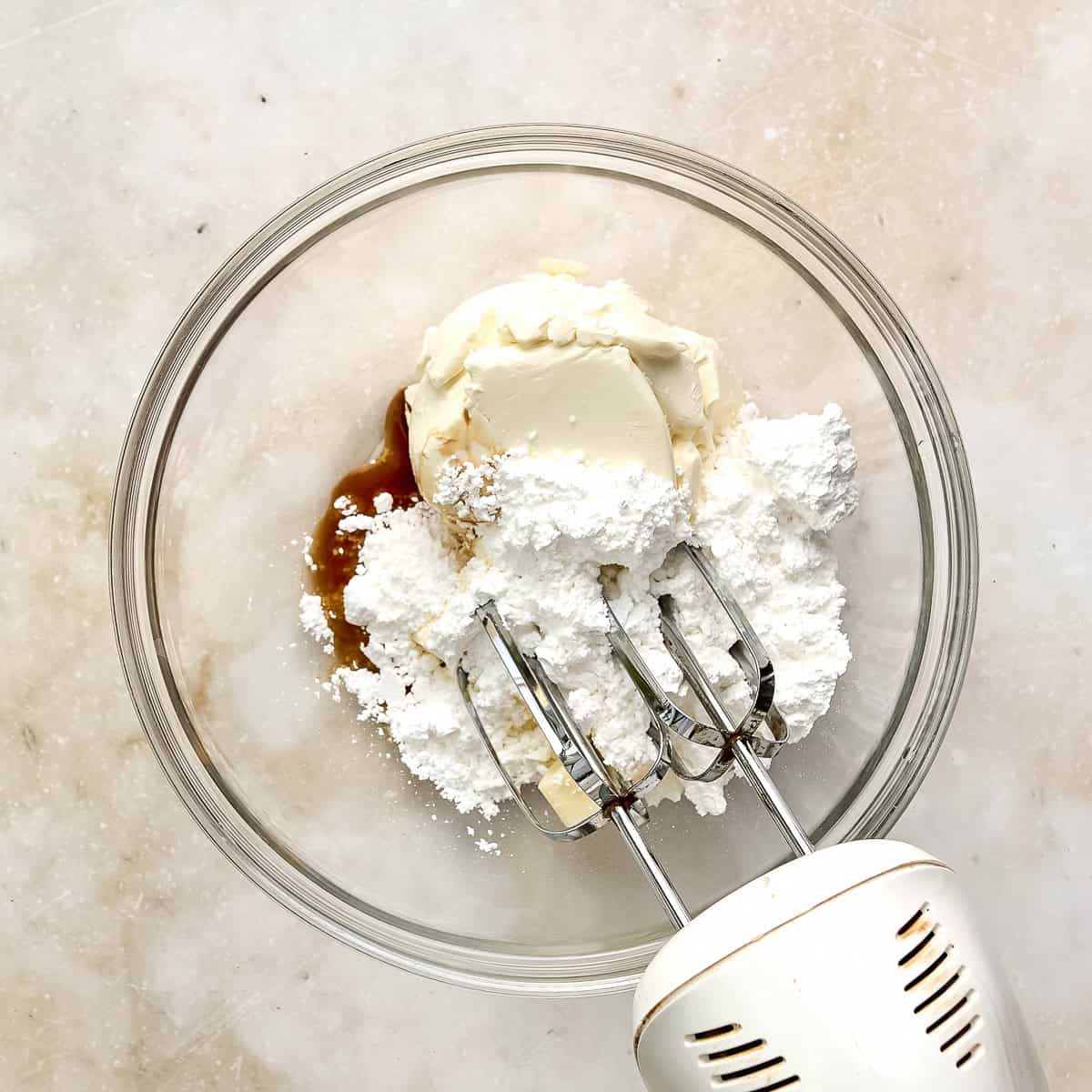 Hand mixer in a bowl with cream cheese, sugar, and vanilla extract.