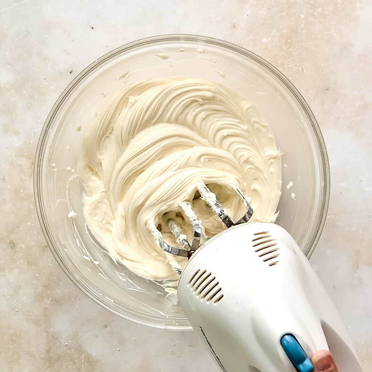 Woman mixing cream cheese frosting in a glass bowl with a hand mixer.