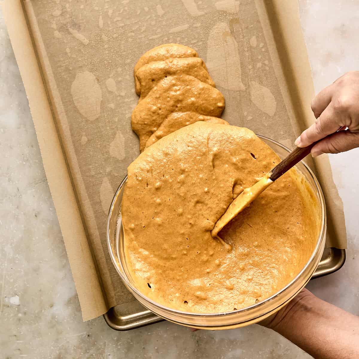 Woman using a rubber spatula to transfer batter to a lined half sheet pan.