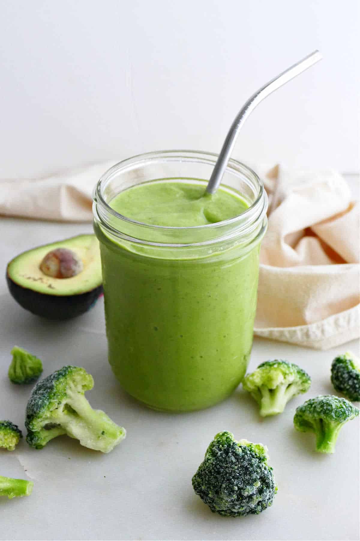 broccoli smoothie in a glass with a steel straw surrounded by ingredients