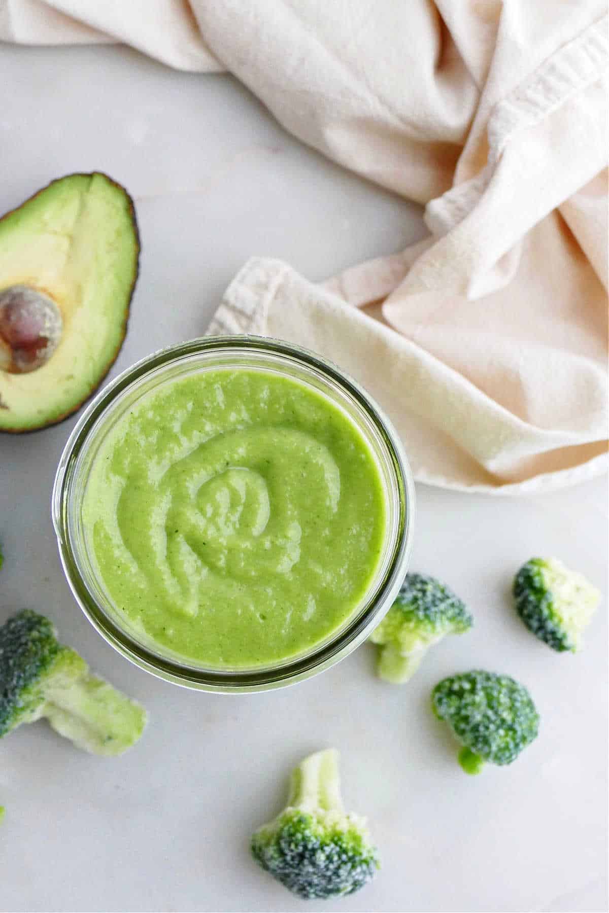 broccoli smoothie in a glass on a counter next to avocado and broccoli