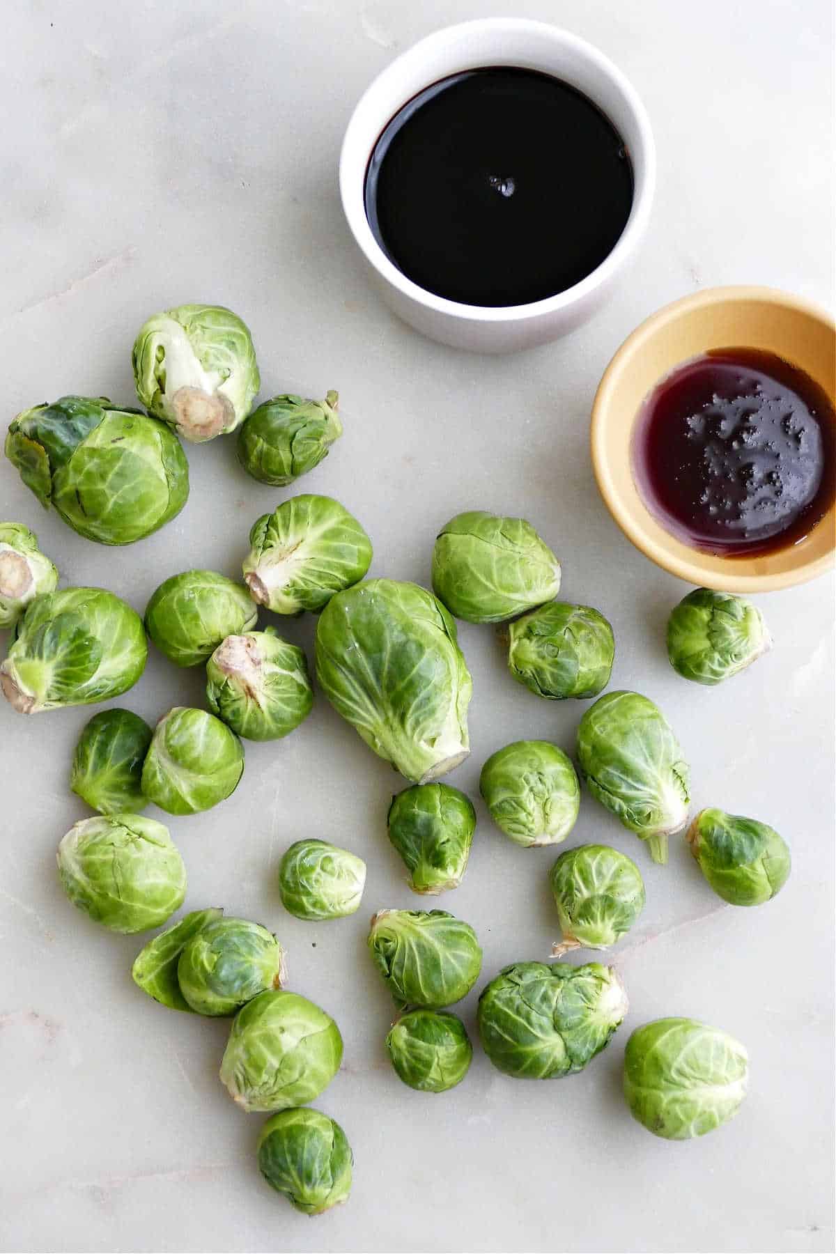 raw Brussels sprouts spread out on a counter next to bowls of maple syrup and balsamic