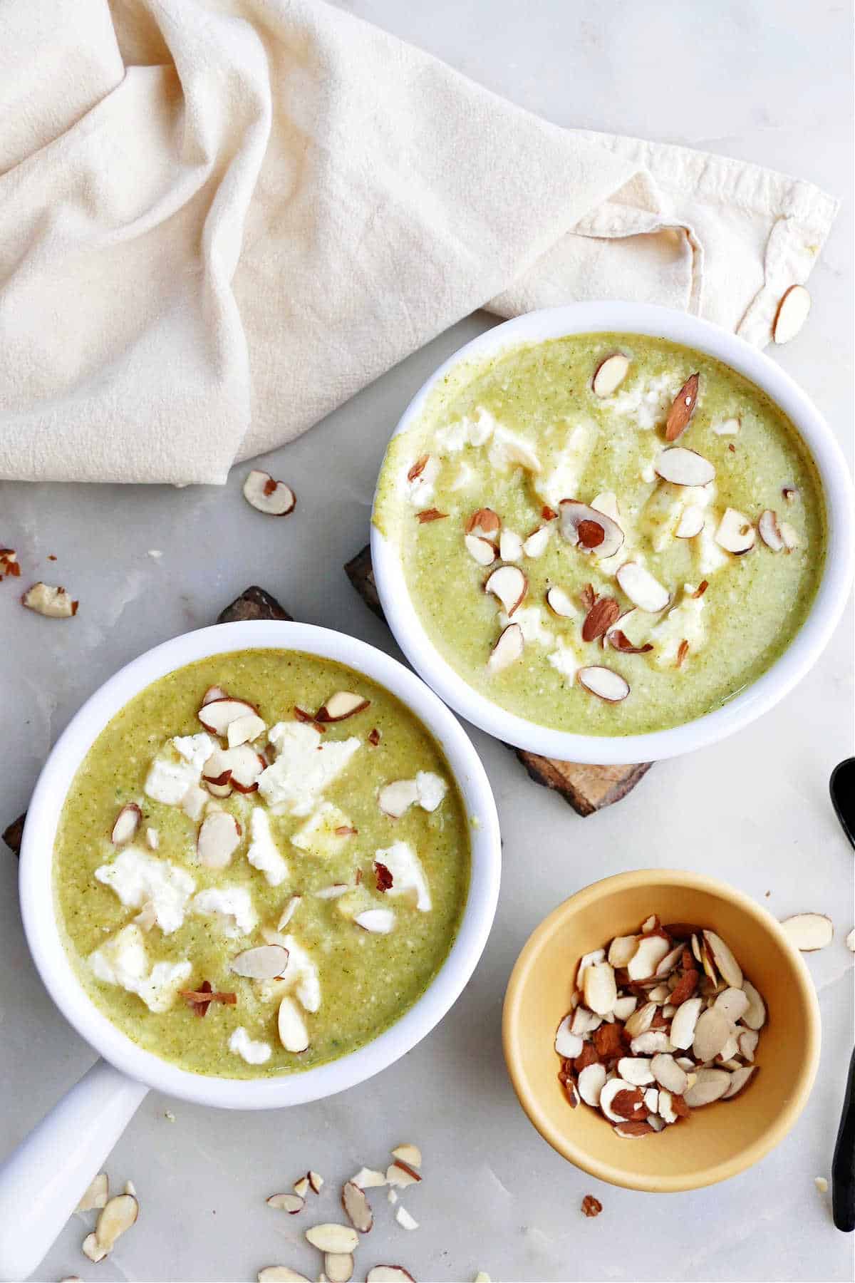 two bowls of broccoli feta soup topped with almonds on a counter