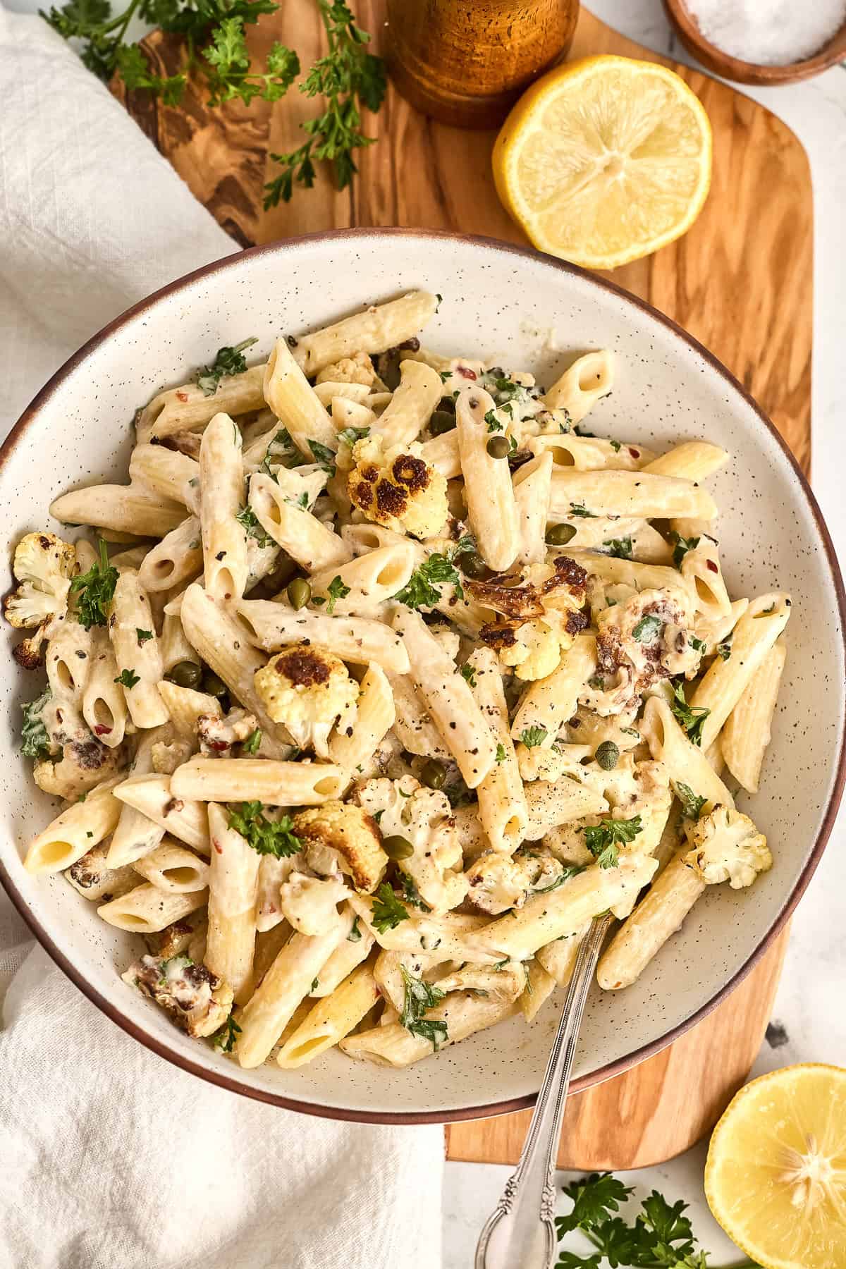 Tahini pasta in a bowl on a board next to lemons, parsley, and a napkin.