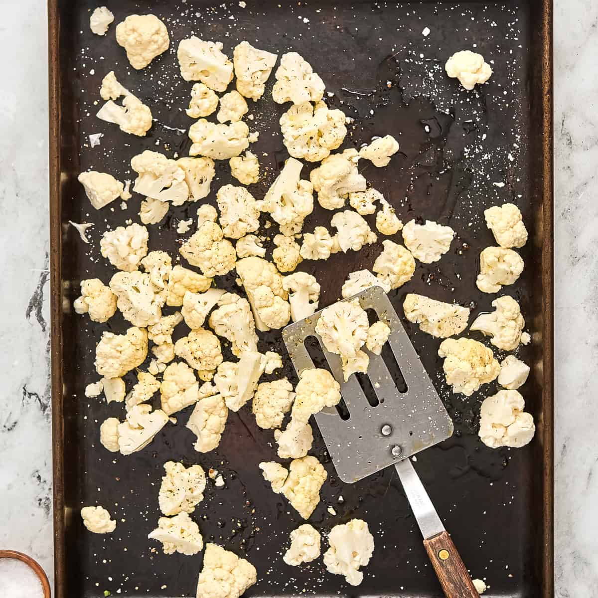 Cauliflower florets on a baking sheet with a spatula before being roasted.