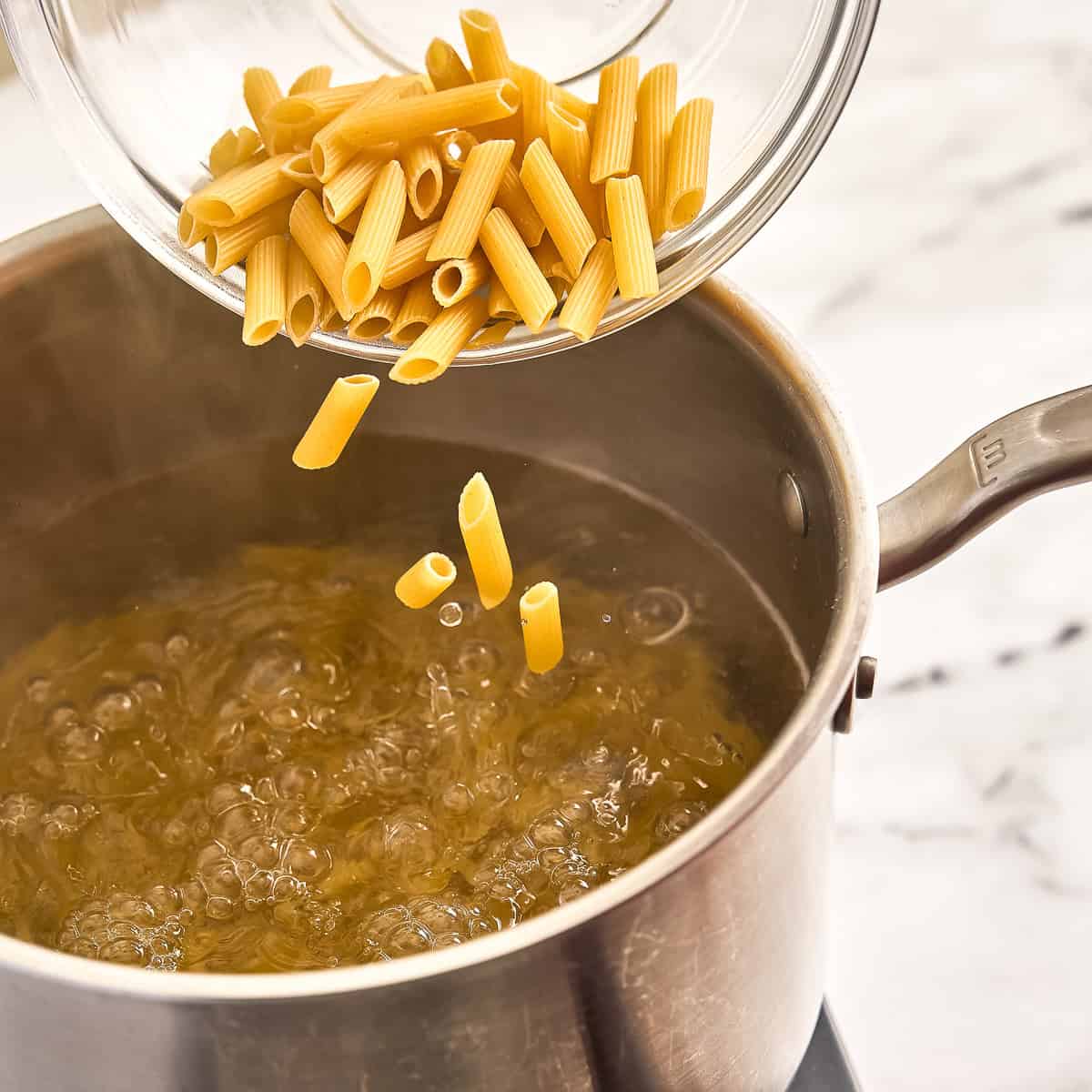 Penne pasta being poured from a bowl into a pot of boiling water.