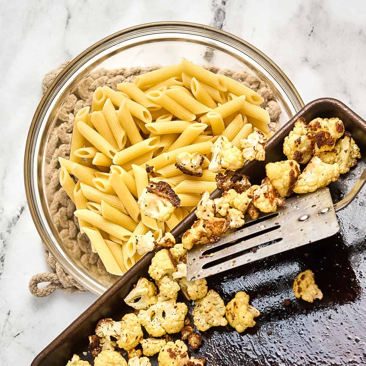 Roasted cauliflower being pushed off a baking sheet onto cooked pasta in a bowl.