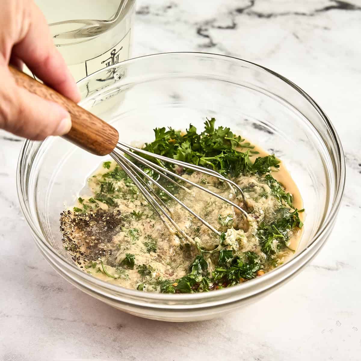 Woman whisking parsley, tahini, lemon juice, garlic, and seasonings in a glass bowl.