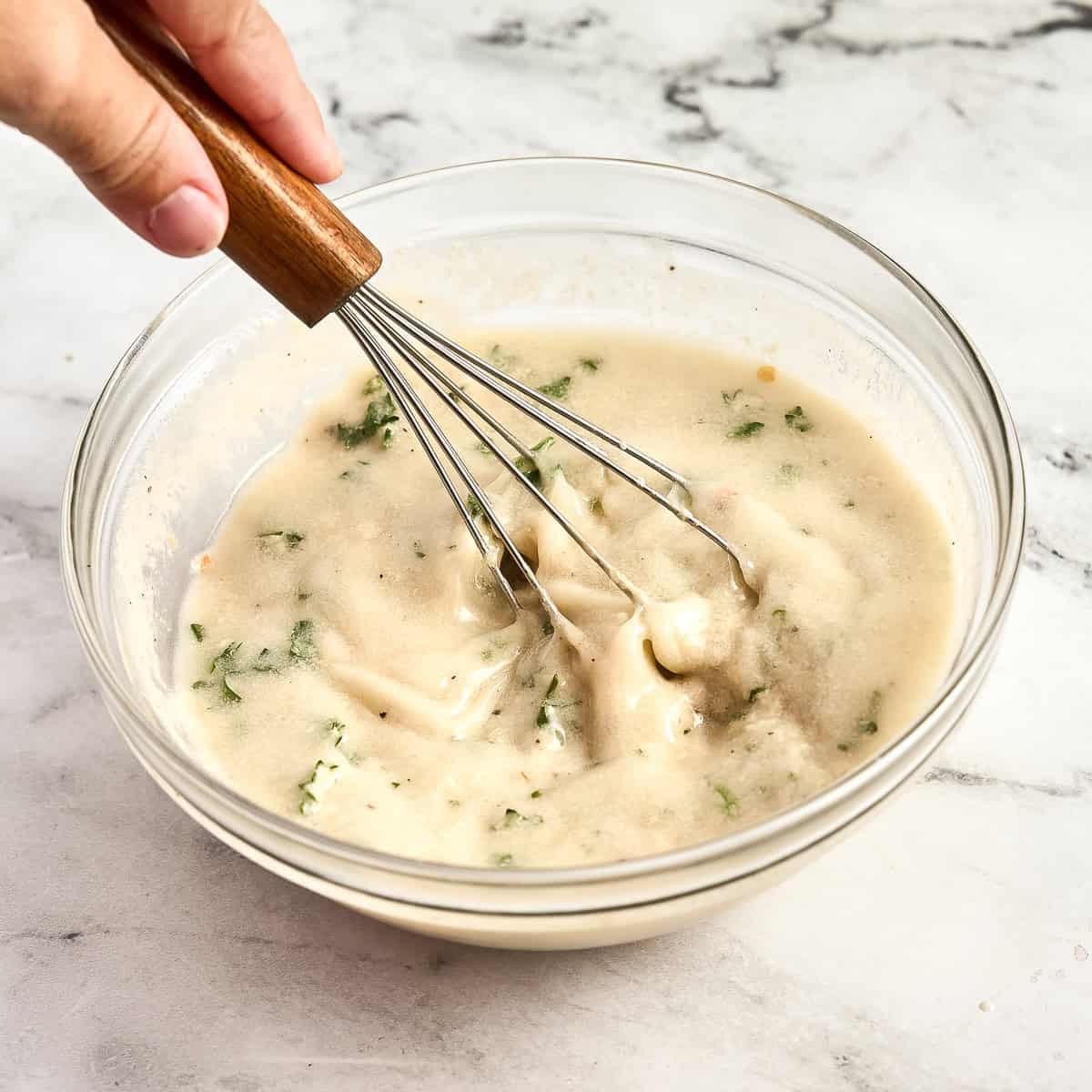 Woman whisking tahini sauce in a glass bowl to pour over pasta.
