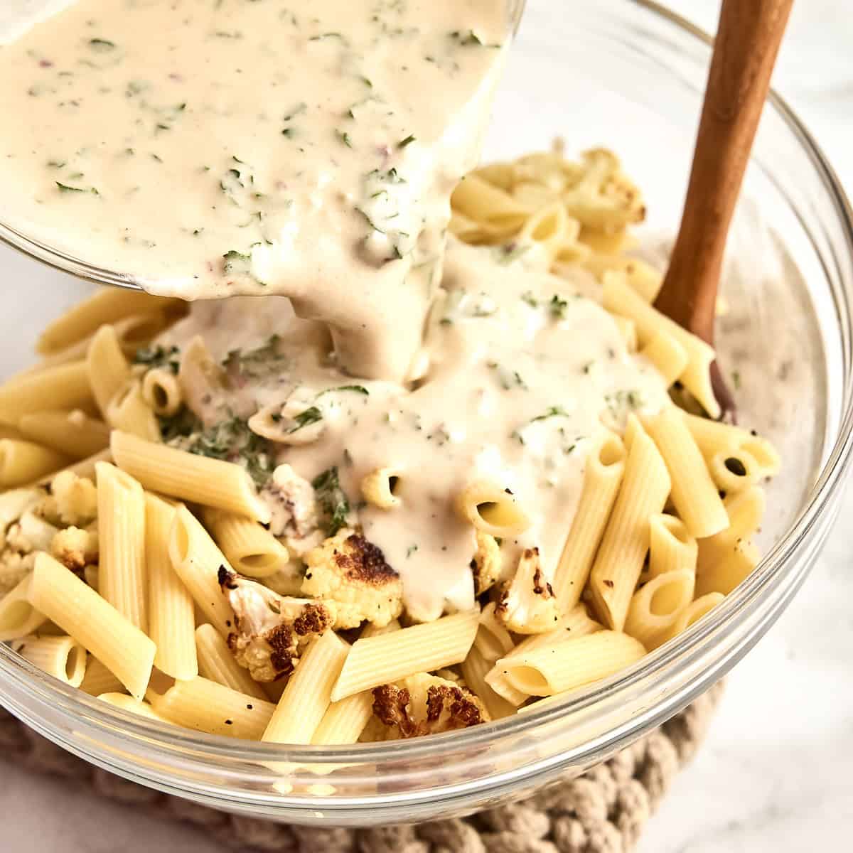 Tahini sauce being poured over cooked penne pasta and cauliflower in a bowl.