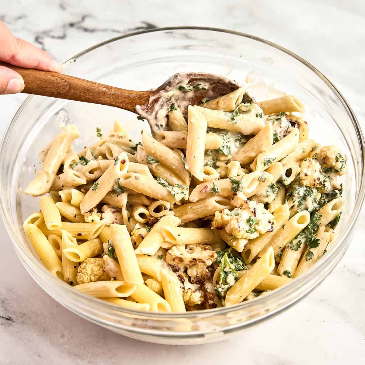 Woman mixing tahini pasta and cauliflower with a wooden spoon in a bowl.