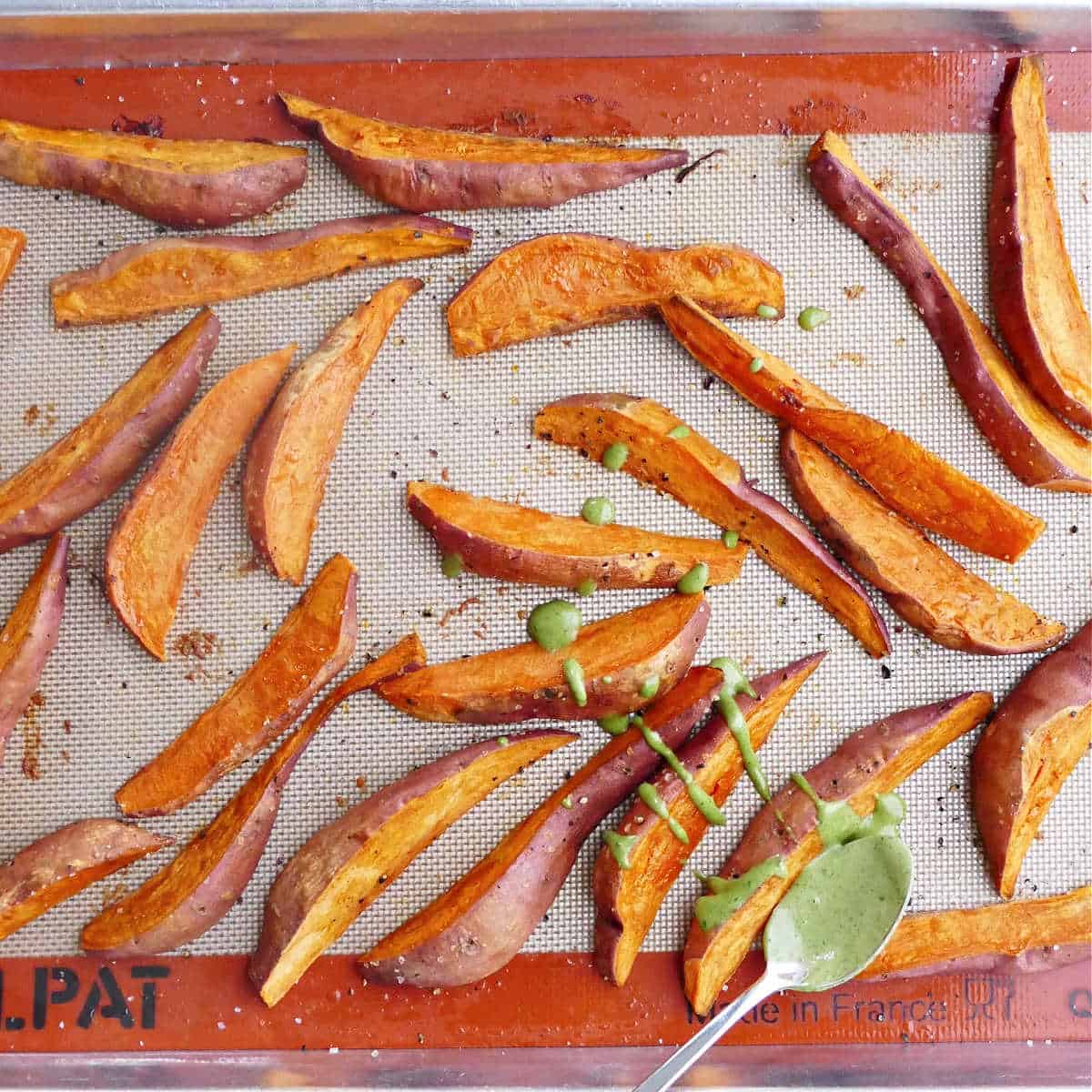 roasted sweet potatoes being drizzled with basil tahini sauce on a baking sheet
