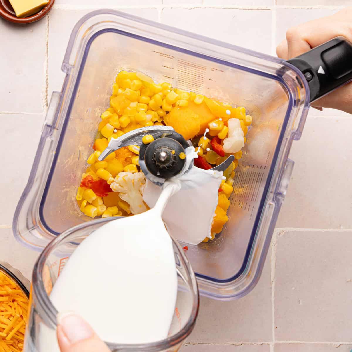 Woman pouring milk into a blender with steamed vegetables to make sauce.