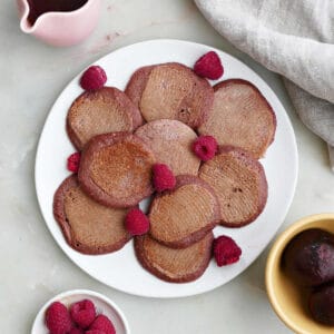 beet pancakes on a plate with raspberries next to toppings on a counter