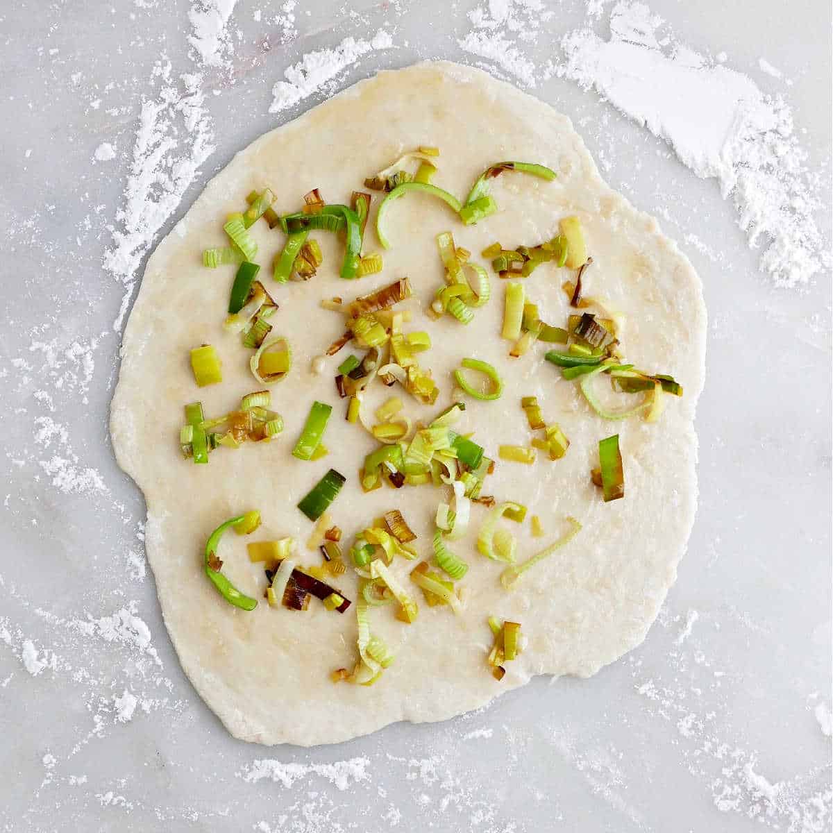 dough rolled into a circle and topped with cooked leeks on a floured surface