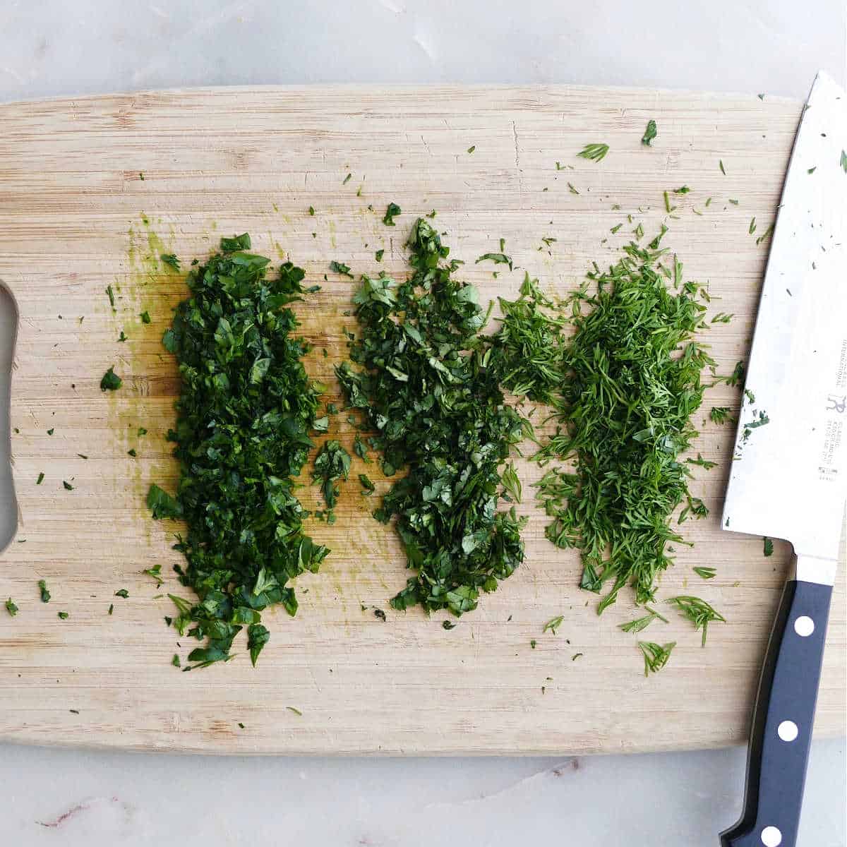 dill, parsley, and cilantro chopped into small pieces on a cutting board with a knife
