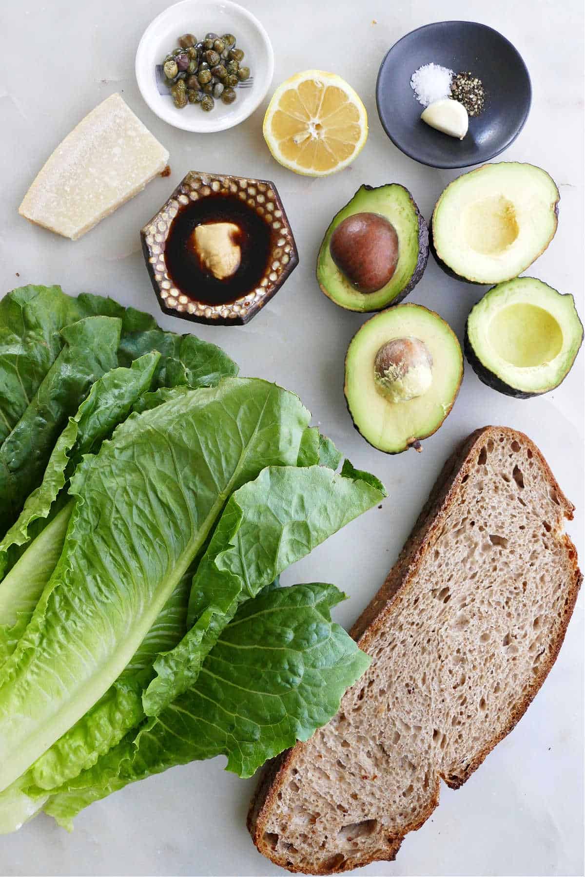 avocados, capers, lemon, cheese, seasonings, romaine, and bread on a counter