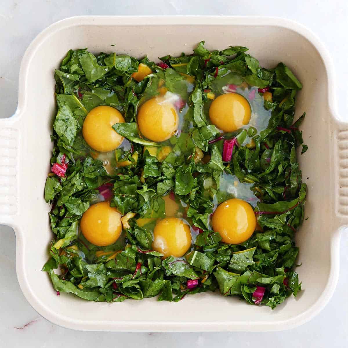 6 eggs cracked into a bed of chard leaves and stems in a baking dish