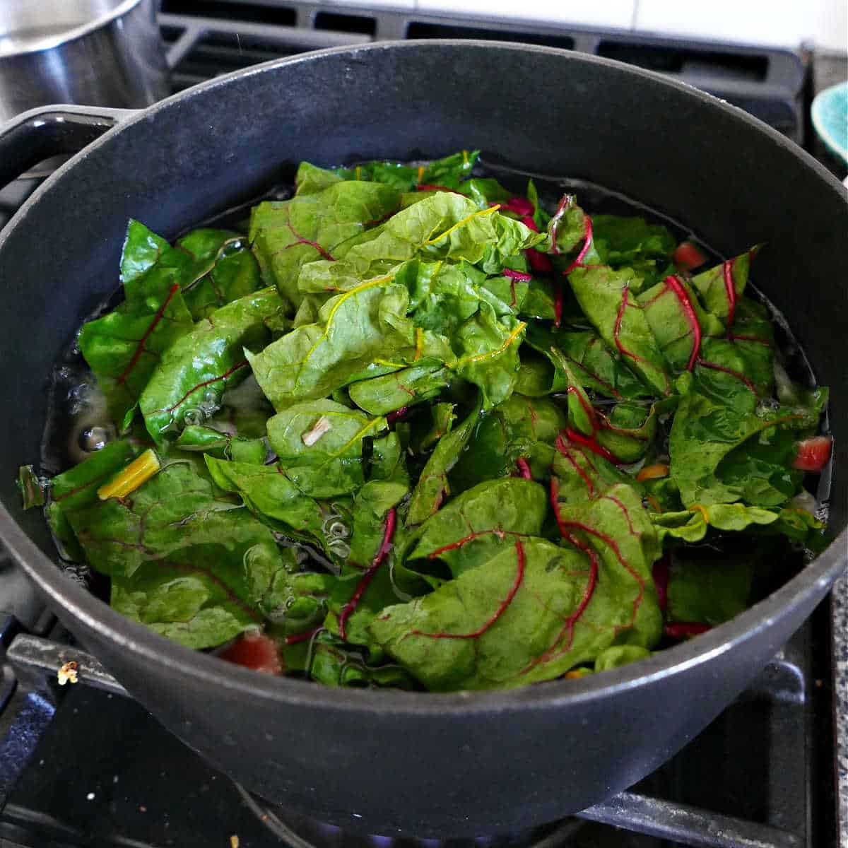 chard leaves being added to a pot of potatoes and chard stems on a stove