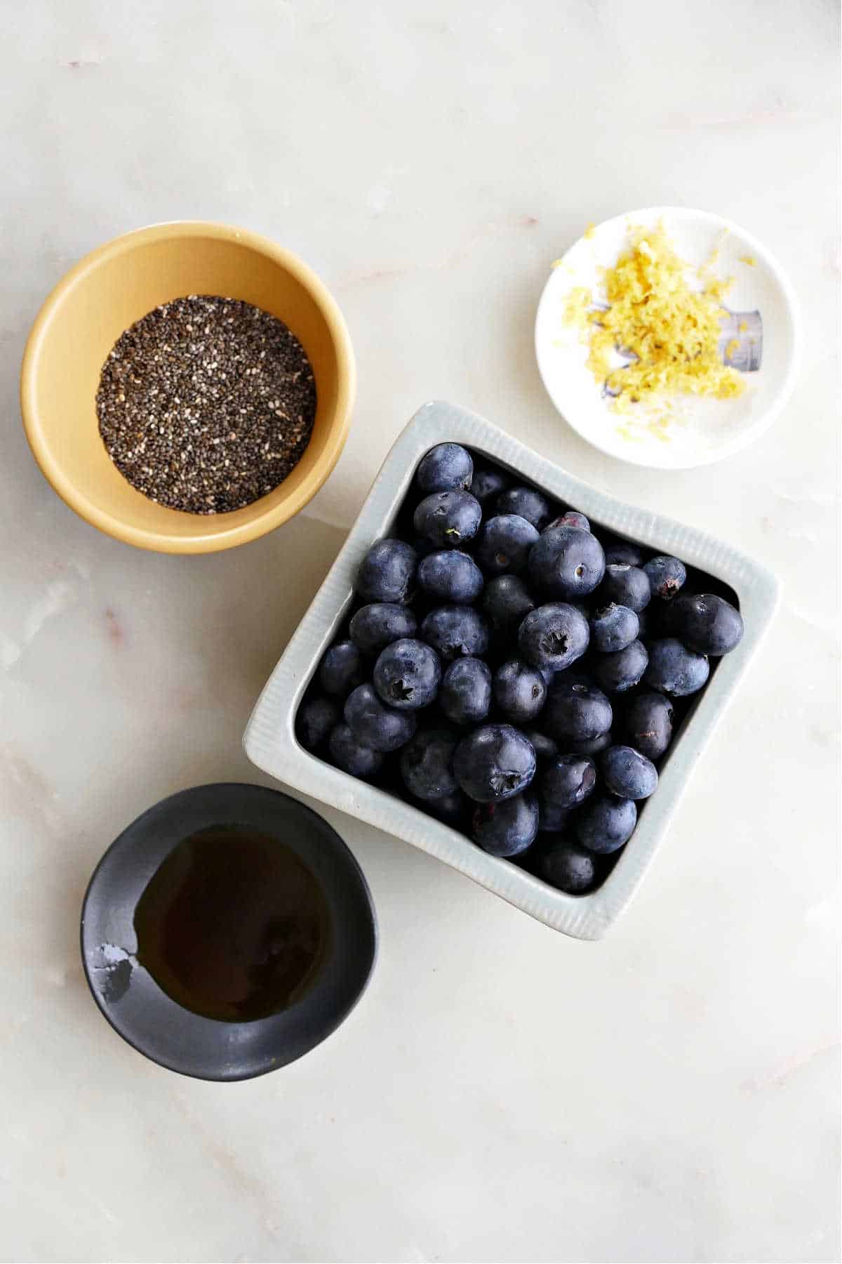 chia seeds, fresh blueberries, lemon zest, and maple syrup on a counter next to each other