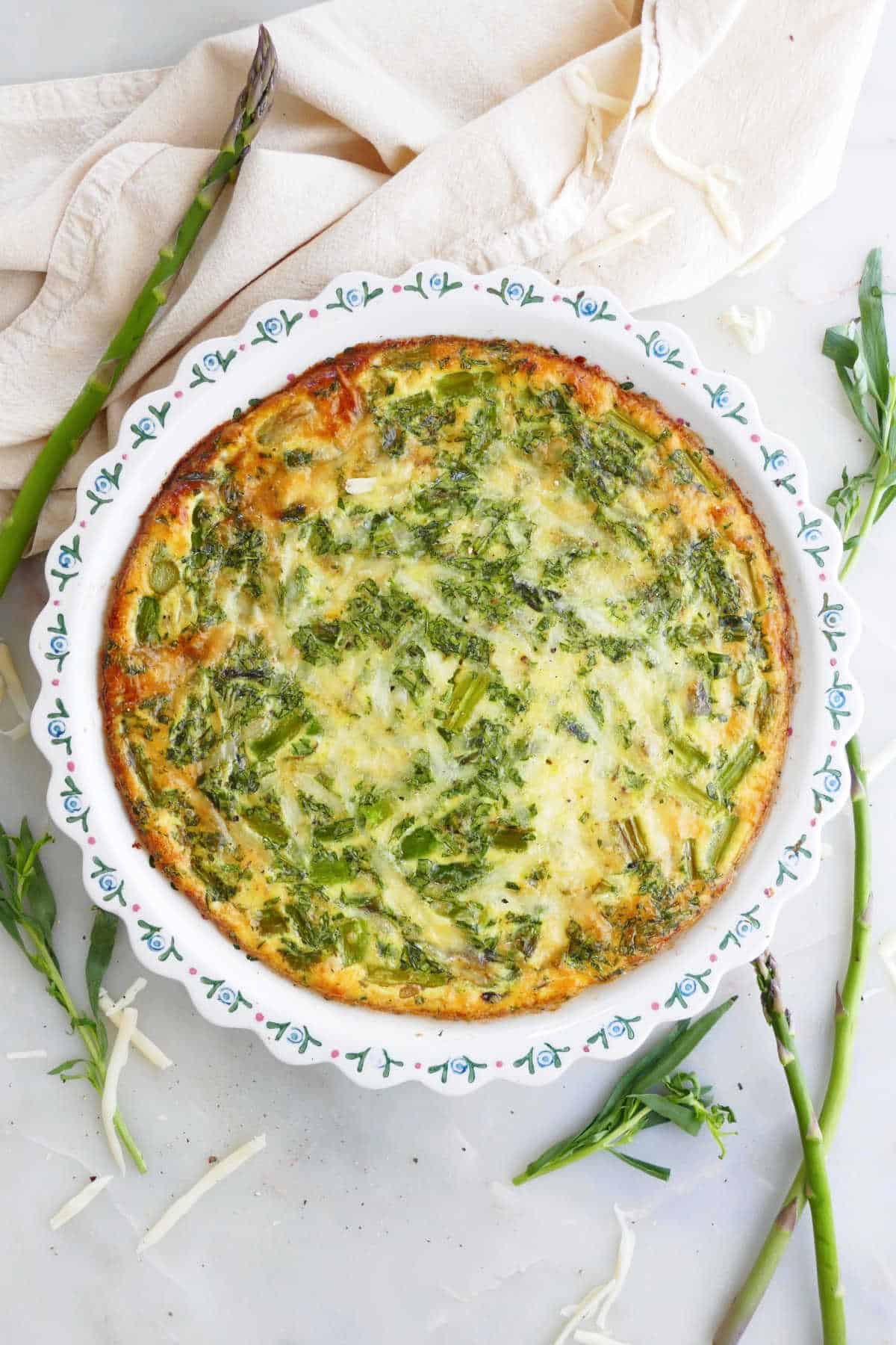 crustless asparagus quiche in a pie plate topped on a counter surrounded by ingredients