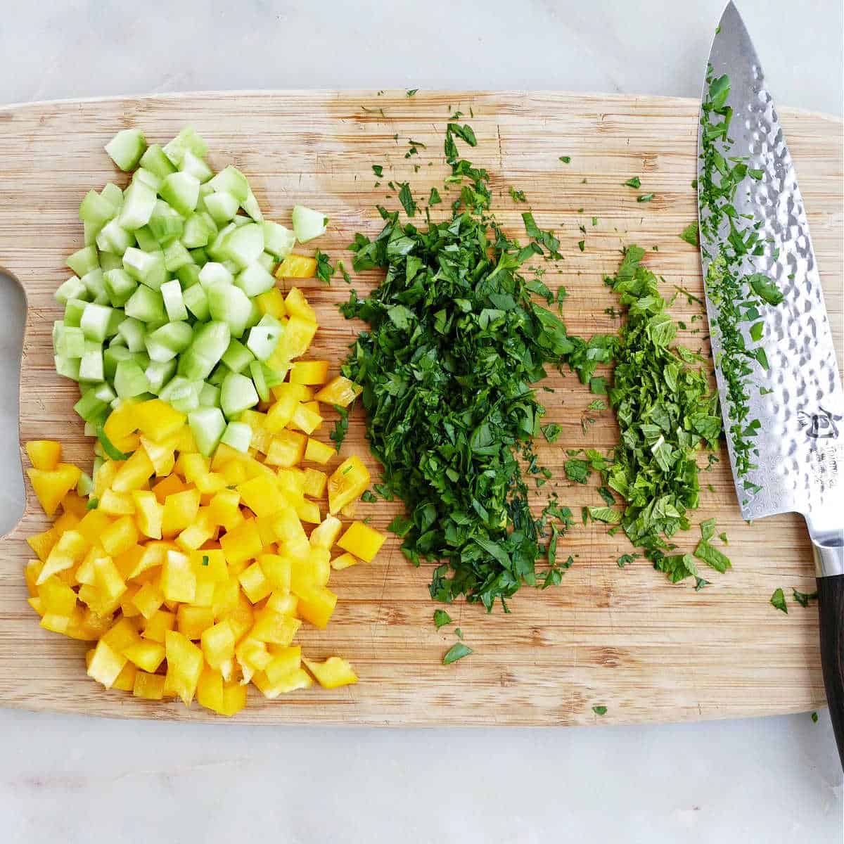 pepper, cucumber, and herbs chopped up on a cutting board with a knife