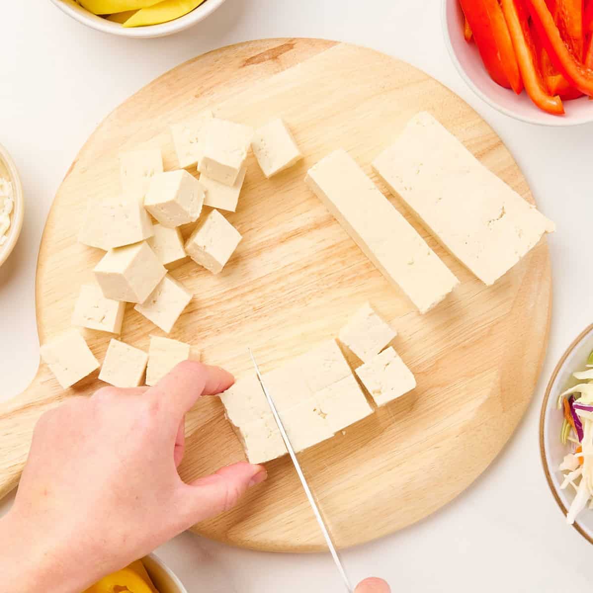 Woman cutting tofu into cubes with a knife on a cutting board.