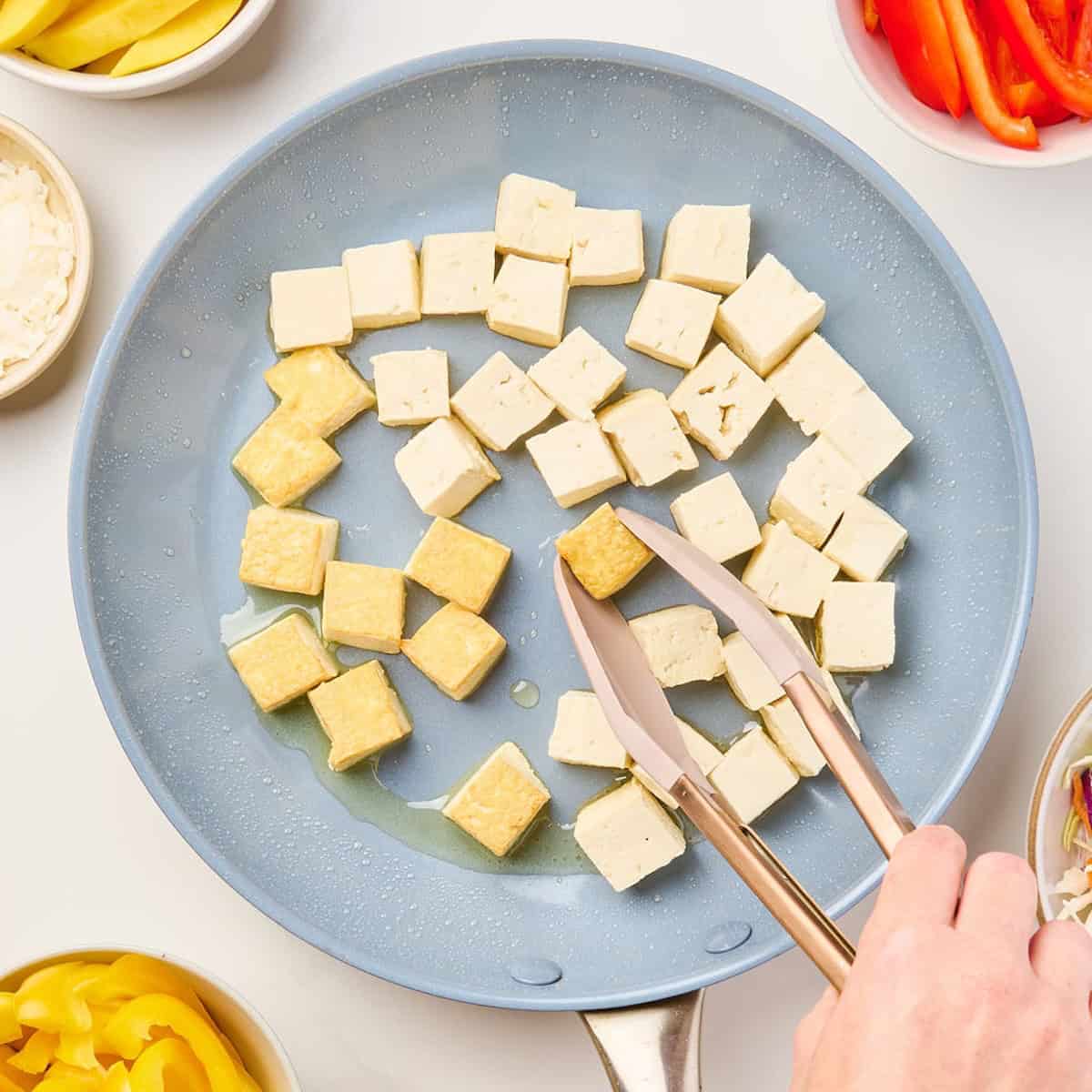 Woman flipping tofu that's frying in a skillet until golden brown.