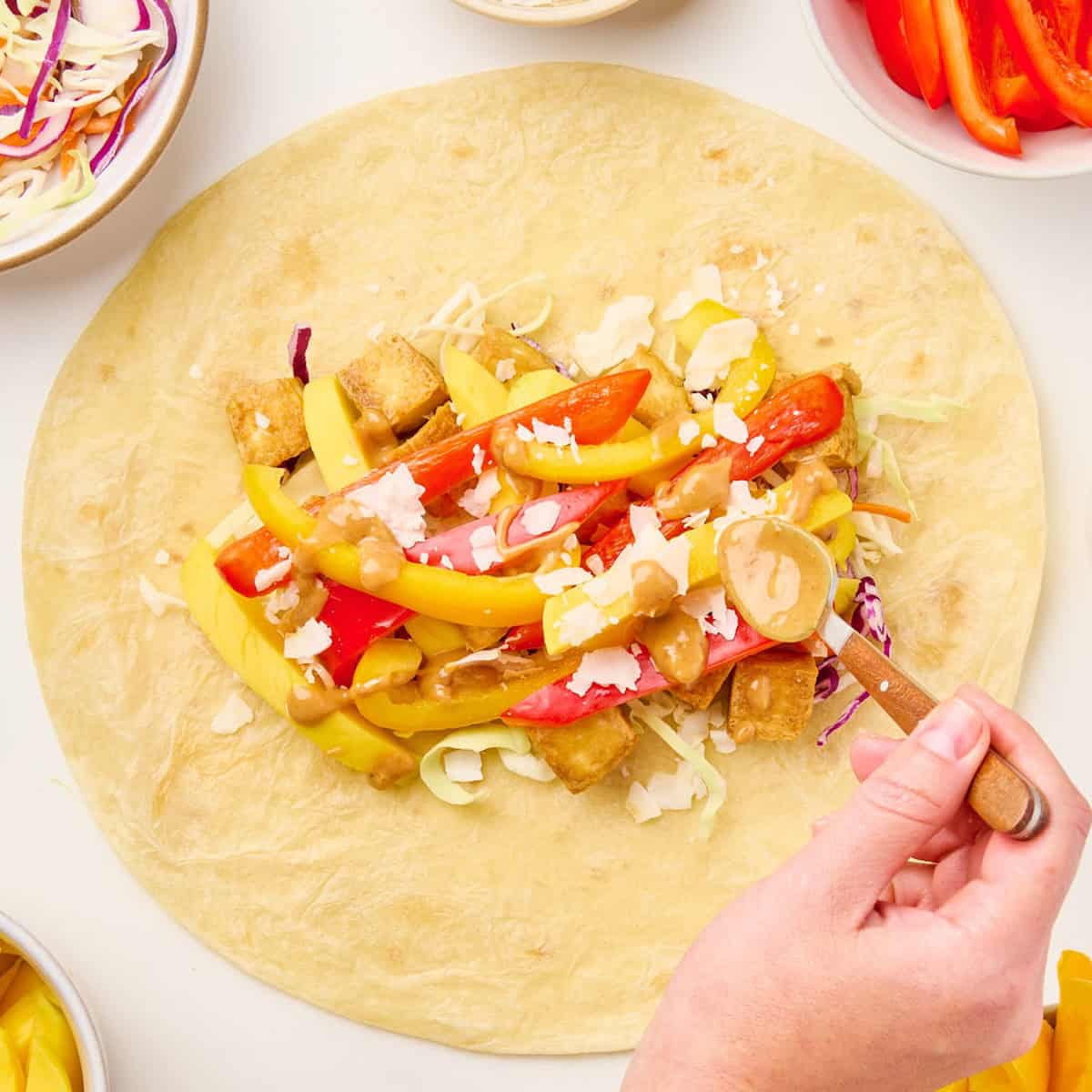 Woman spooning peanut sauce over ingredients for tofu wraps in a tortilla.
