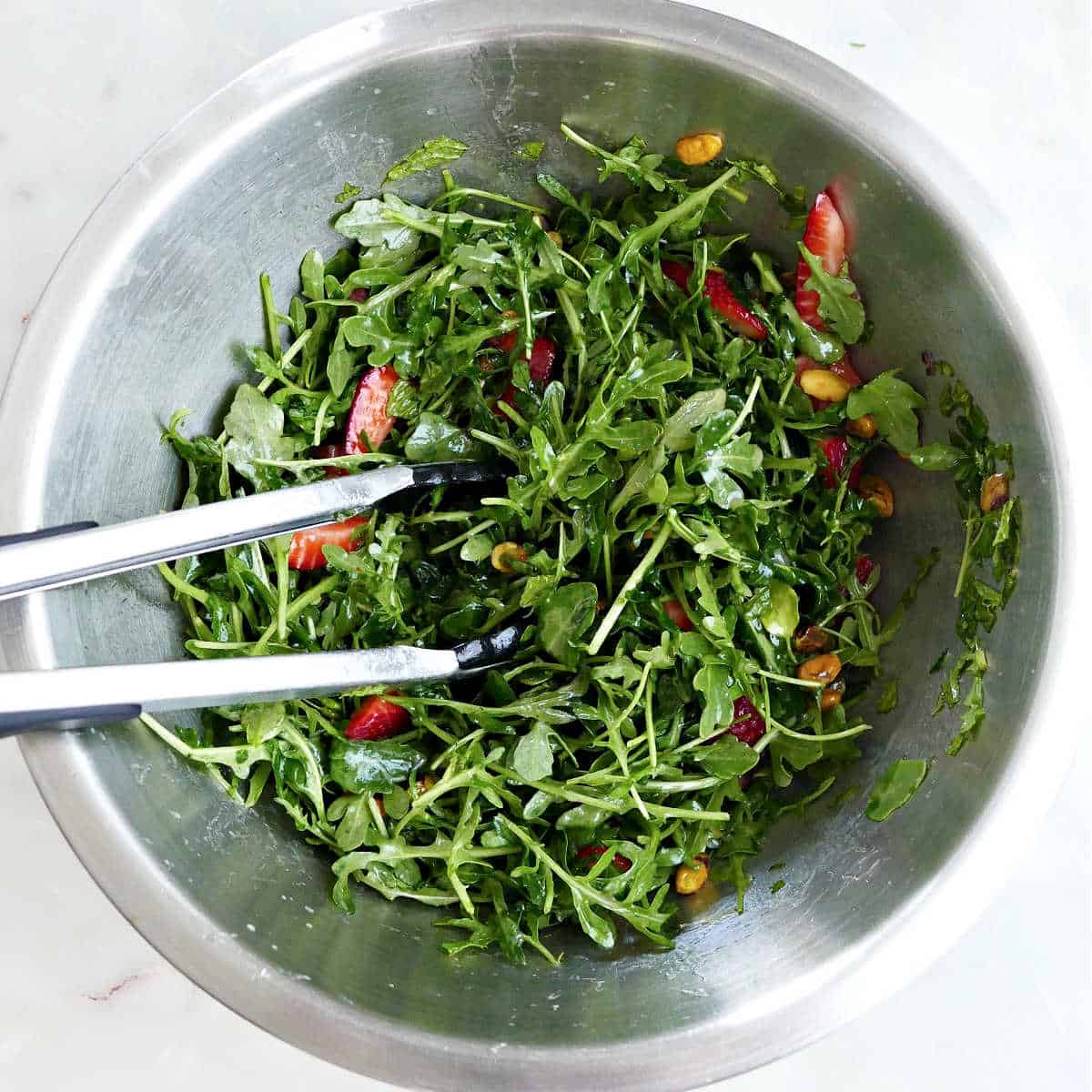 ingredients for arugula salad in a mixing bowl being tossed with tongs