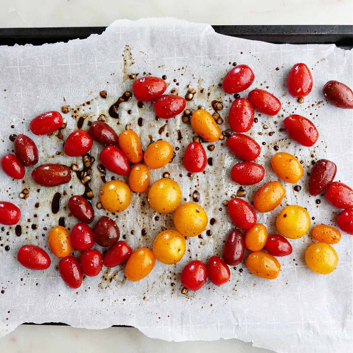 cherry tomatoes tossed with balsamic, garlic, and oil on a lined baking sheet