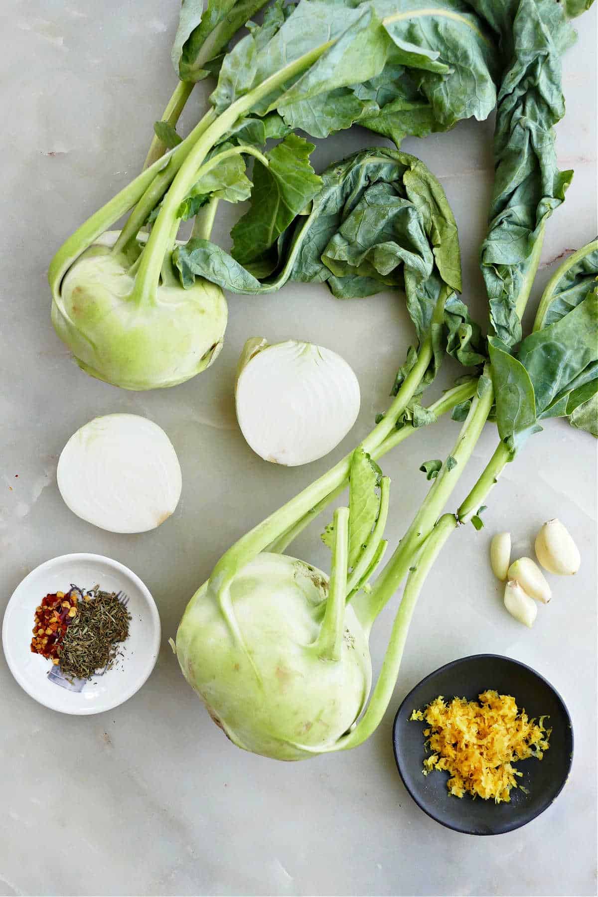 two kohlrabi bulbs with leaves, onion, garlic, lemon zest, and spices on a counter