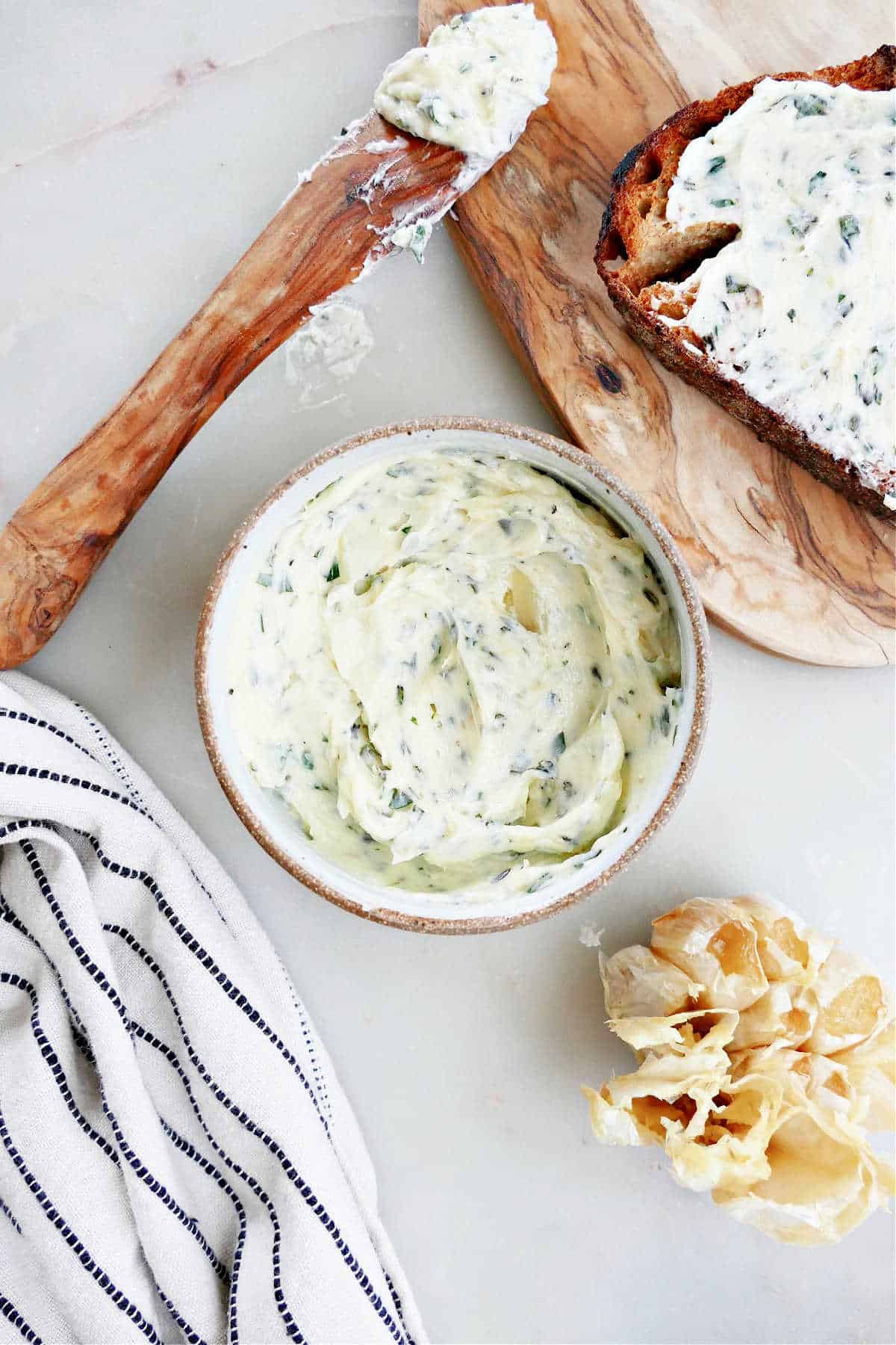 garlic herb butter in a bowl next to roasted garlic, bread, a knife, and napkin