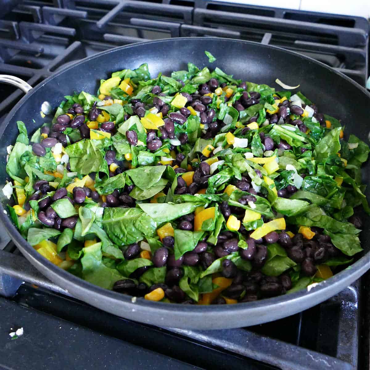 spinach, beans, peppers, onions, and corn cooking in a skillet on a stove