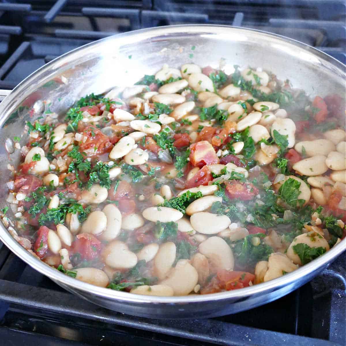 gigante beans, tomatoes, kale, and other seasonings simmering in a skillet on the stove