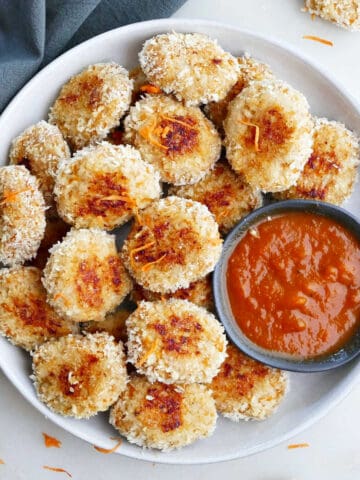 Chicken nuggets with veggies on a white plate next to a bowl of red sauce.